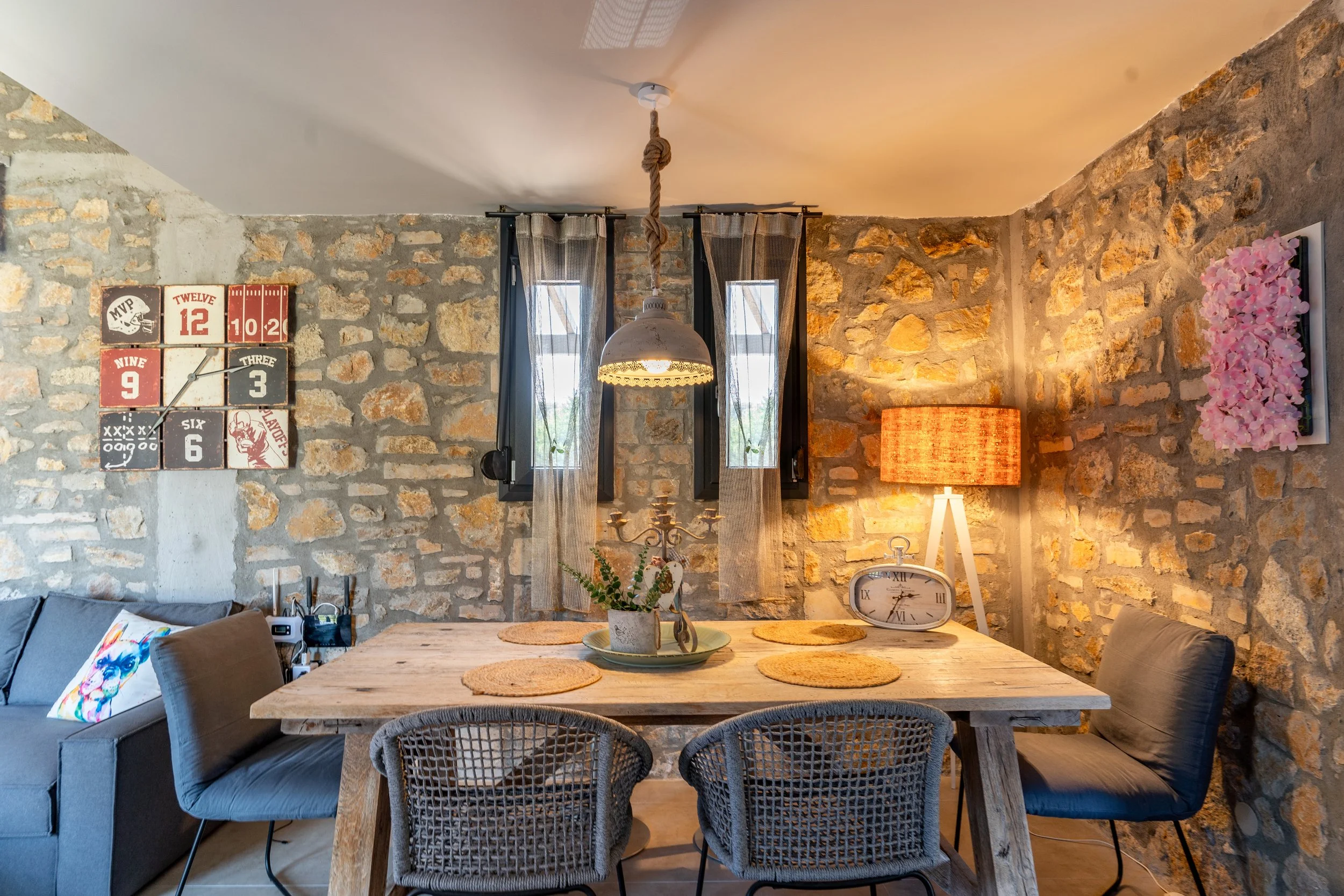 Dining room with a rustic stone wall, a wooden table with six woven placemats, a floral centerpiece, and a vintage clock. There are two black-framed windows, a hanging lamp, a pink floral wall art, and a blue sofa with a colorful pillow.