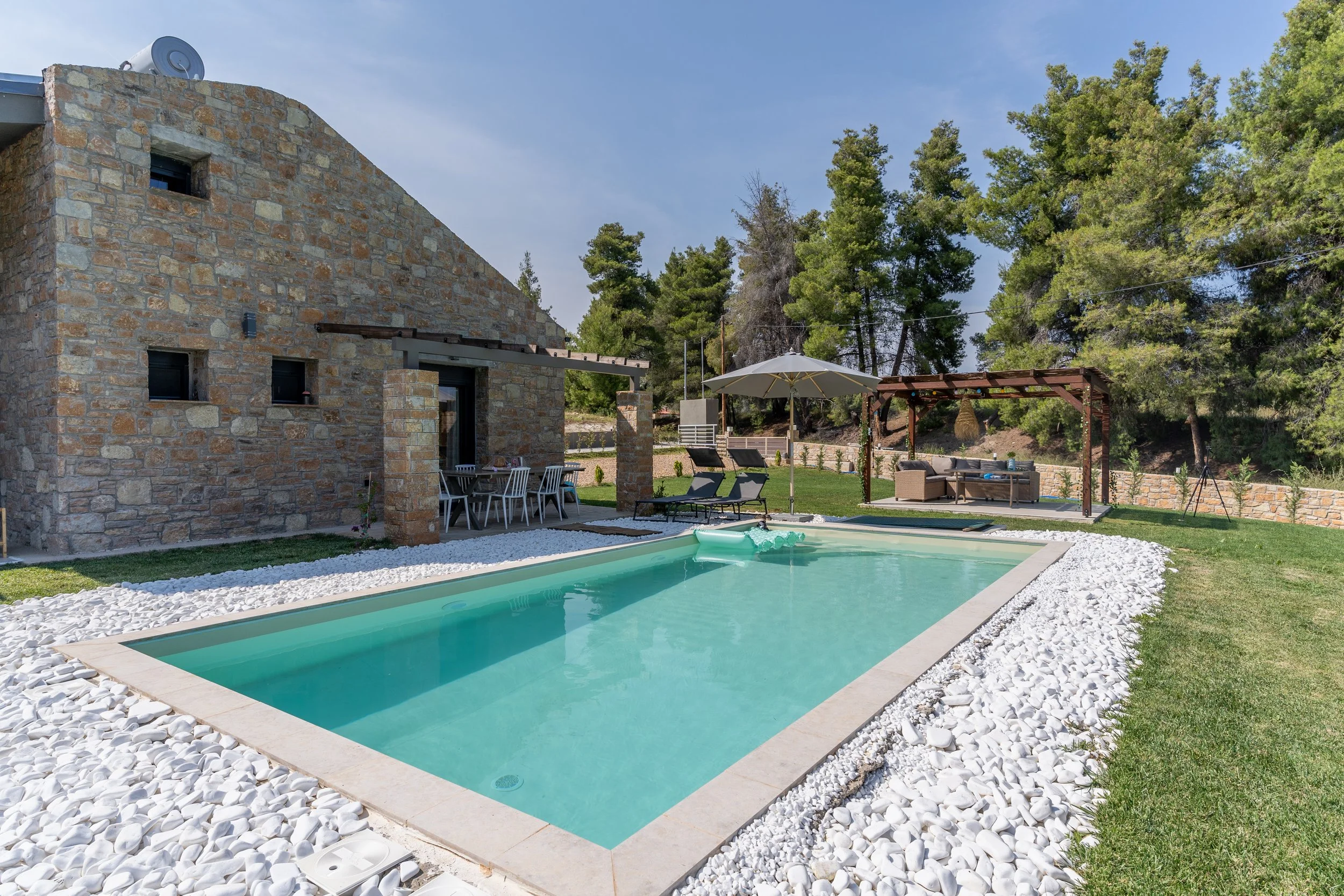 Residential backyard with a rectangular swimming pool surrounded by white rocks and green grass, a stone house, outdoor dining table, lounge chairs with umbrella, and patio seating with a pergola, all set against a backdrop of trees.