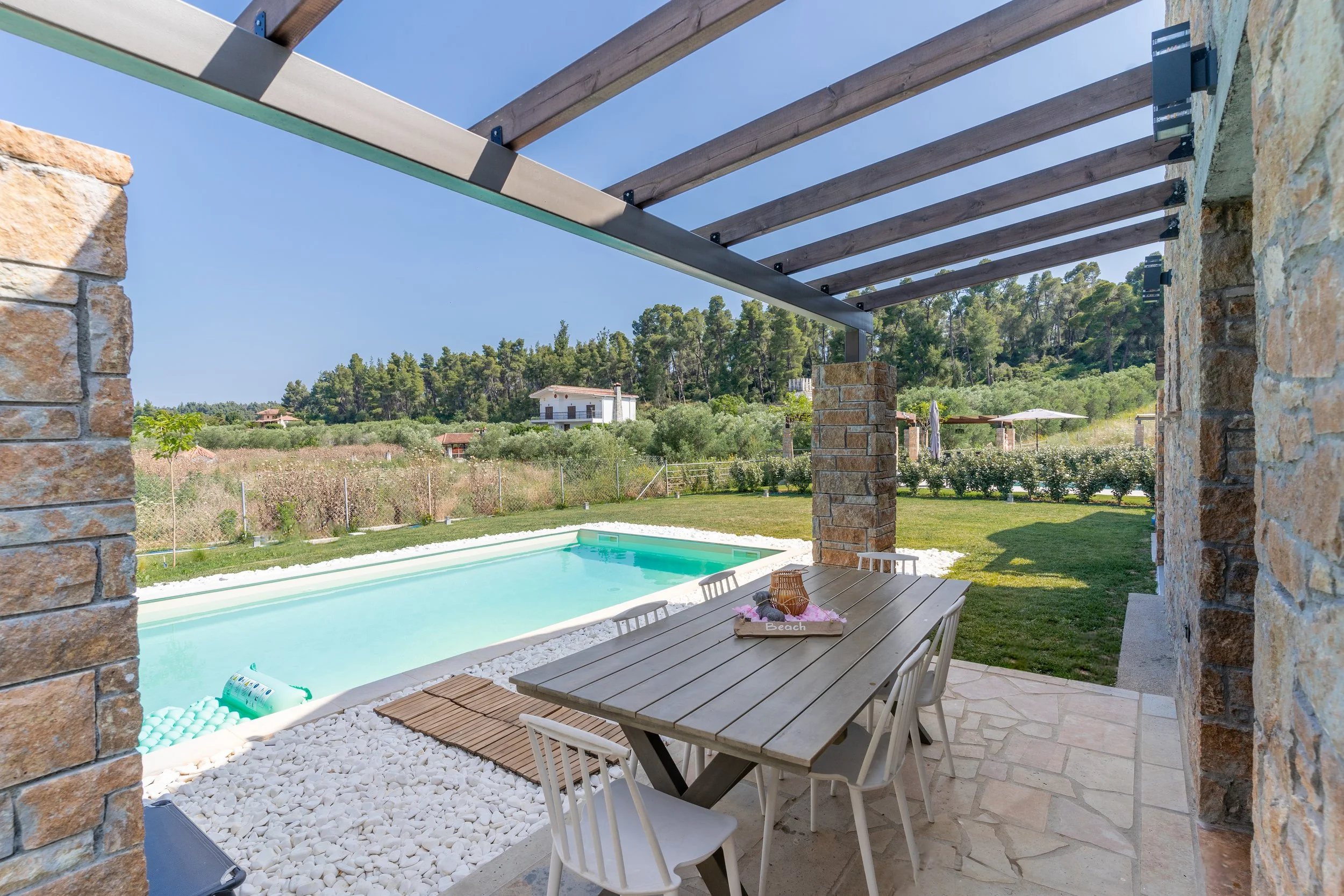 Backyard patio with outdoor dining table, chairs, and swimming pool under a pergola with stone and wood columns, overlooking a green lawn and trees.