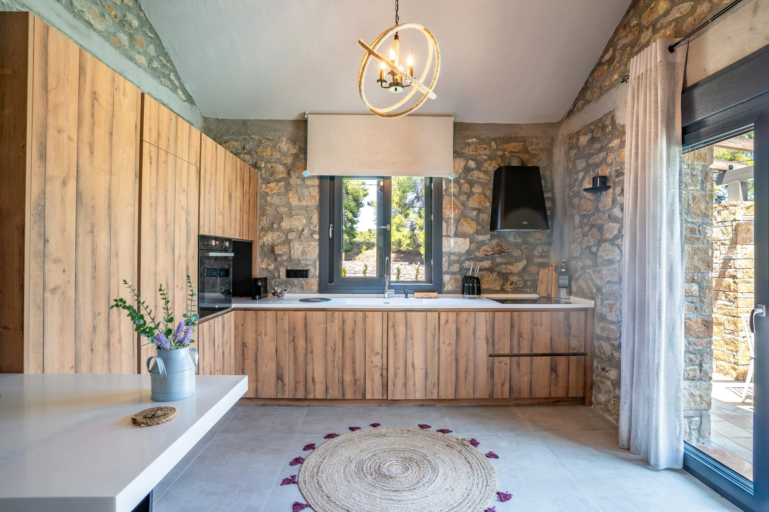Kitchen with wooden cabinets, stone walls, a window above the sink, and a sliding glass door with curtains. There is a white countertop with small appliances, a vase with purple flowers, and a round woven rug on the tile floor.