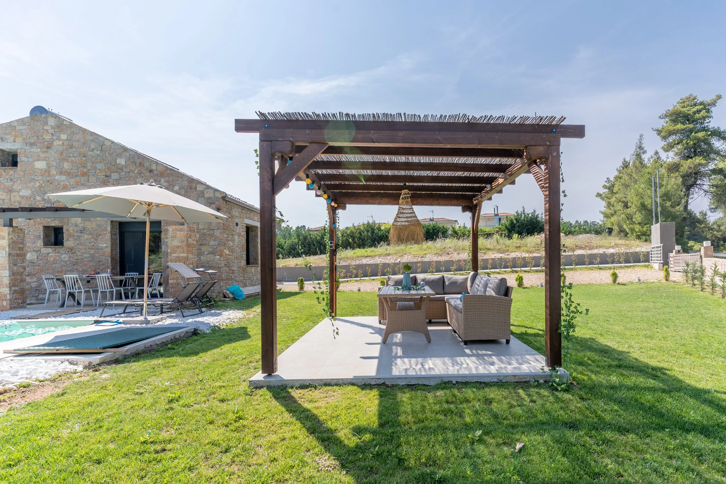 Outdoor patio with a wooden pergola, woven chairs, and a small table in a green backyard with a house and trees in the background.