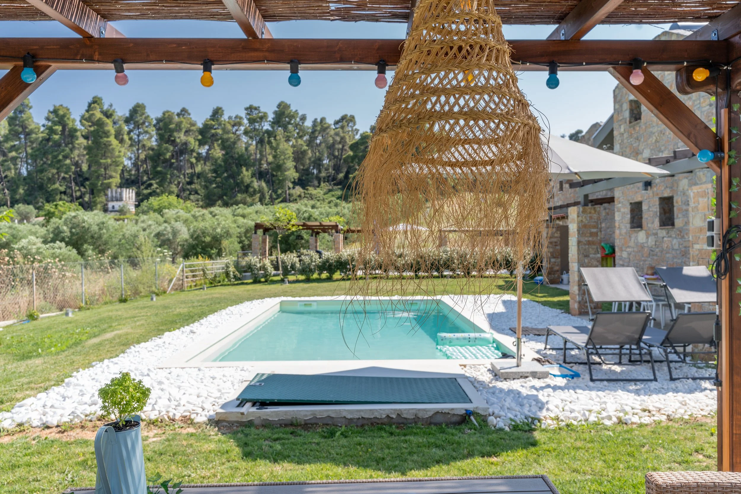 Backyard with swimming pool, lounge chairs, umbrella, decorative hanging light, and surrounding greenery.