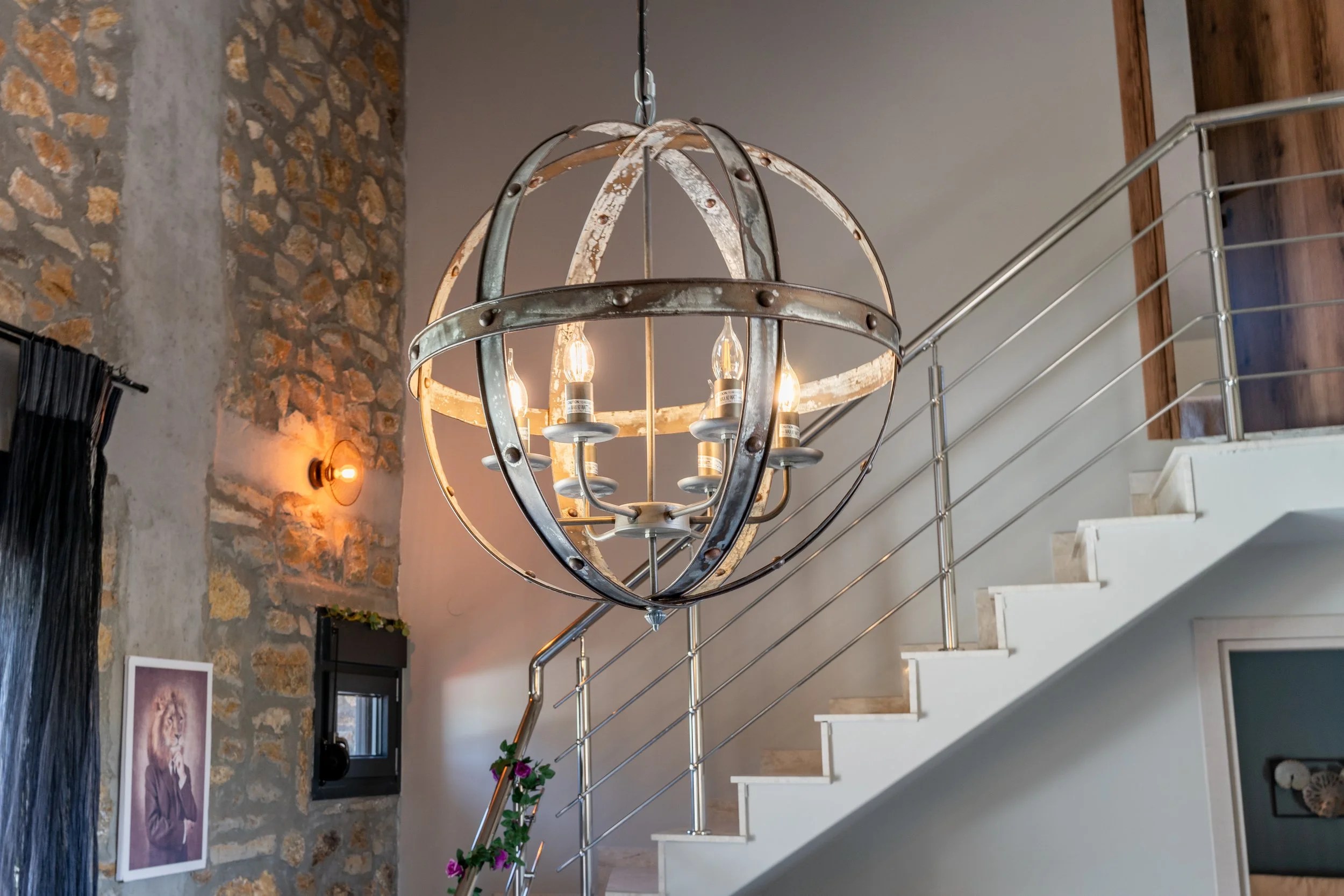 A rustic metal chandelier hangs from the ceiling of a modern interior space, featuring a spherical design with exposed light bulbs inside. The background includes a stone accent wall, a wall-mounted light fixture, and a staircase with a metal railing