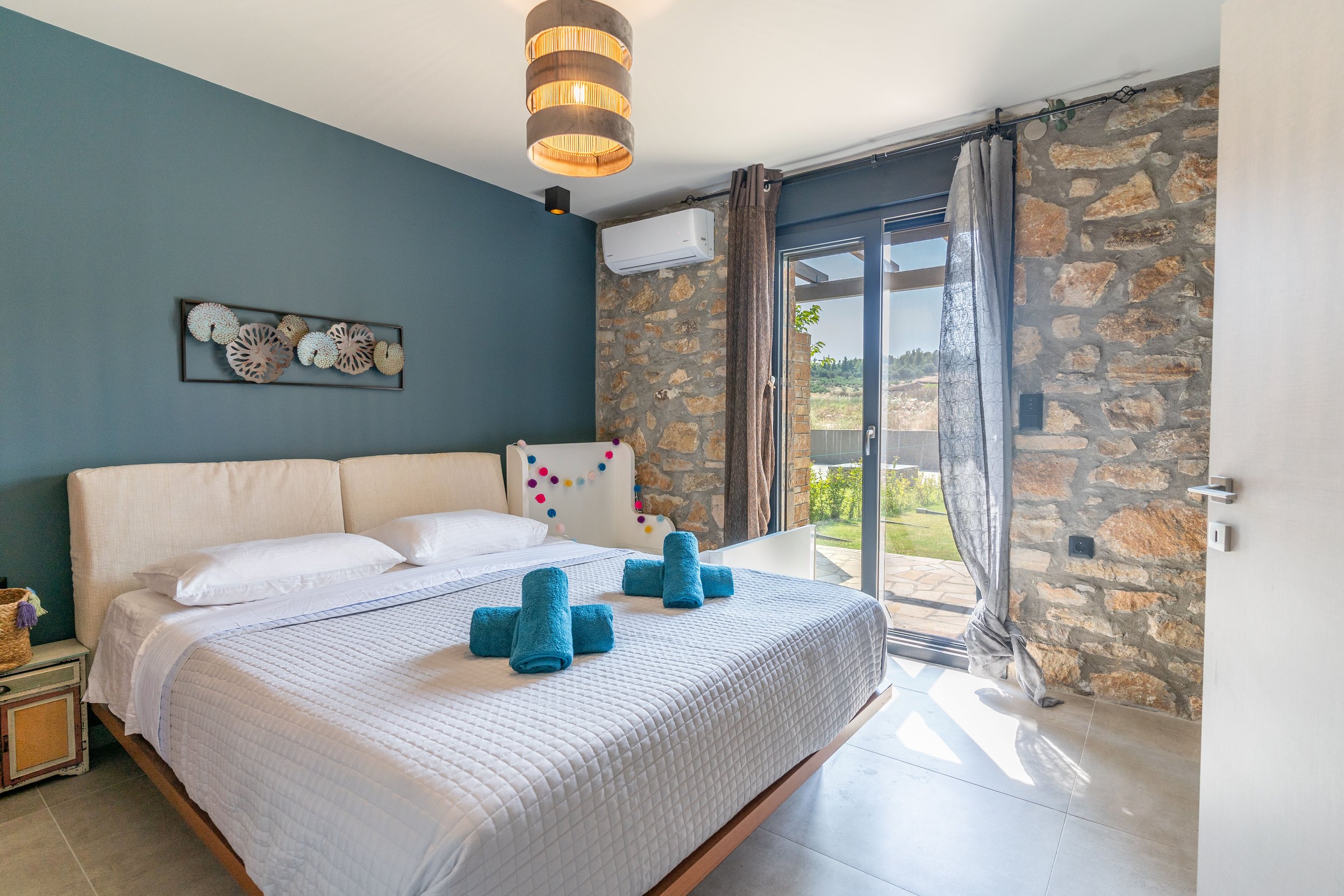 A modern bedroom with a large bed, decorated with two turquoise towels folded at the foot, and a stone accent wall with a sliding glass door leading outside. The room features a blue accent wall, artwork of seashells above the bed, and a small side table. Sunlight streams through the curtains onto a patio area.