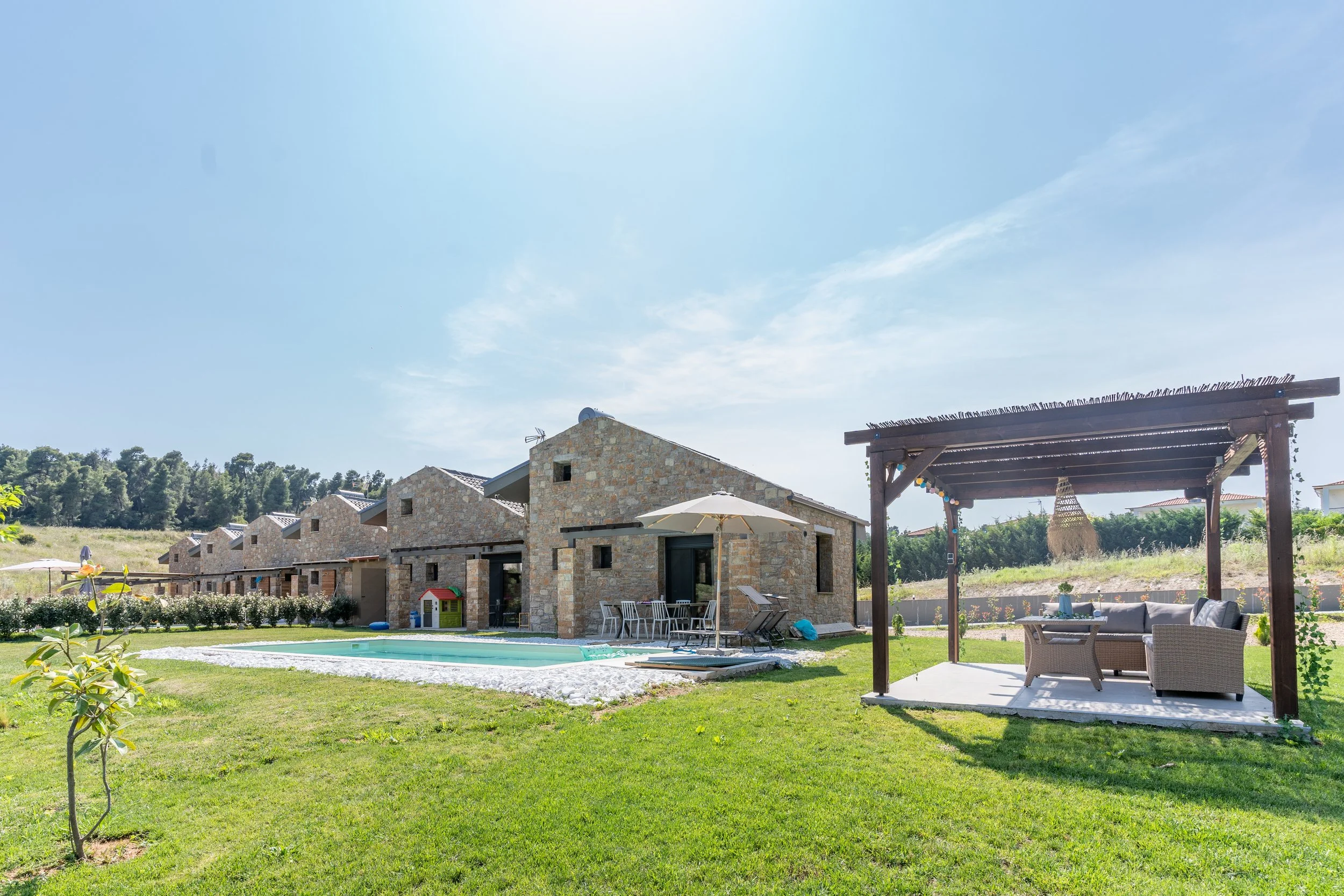 A backyard with a swimming pool, stone houses, and a wooden pergola with outdoor furniture under a sunny sky.
