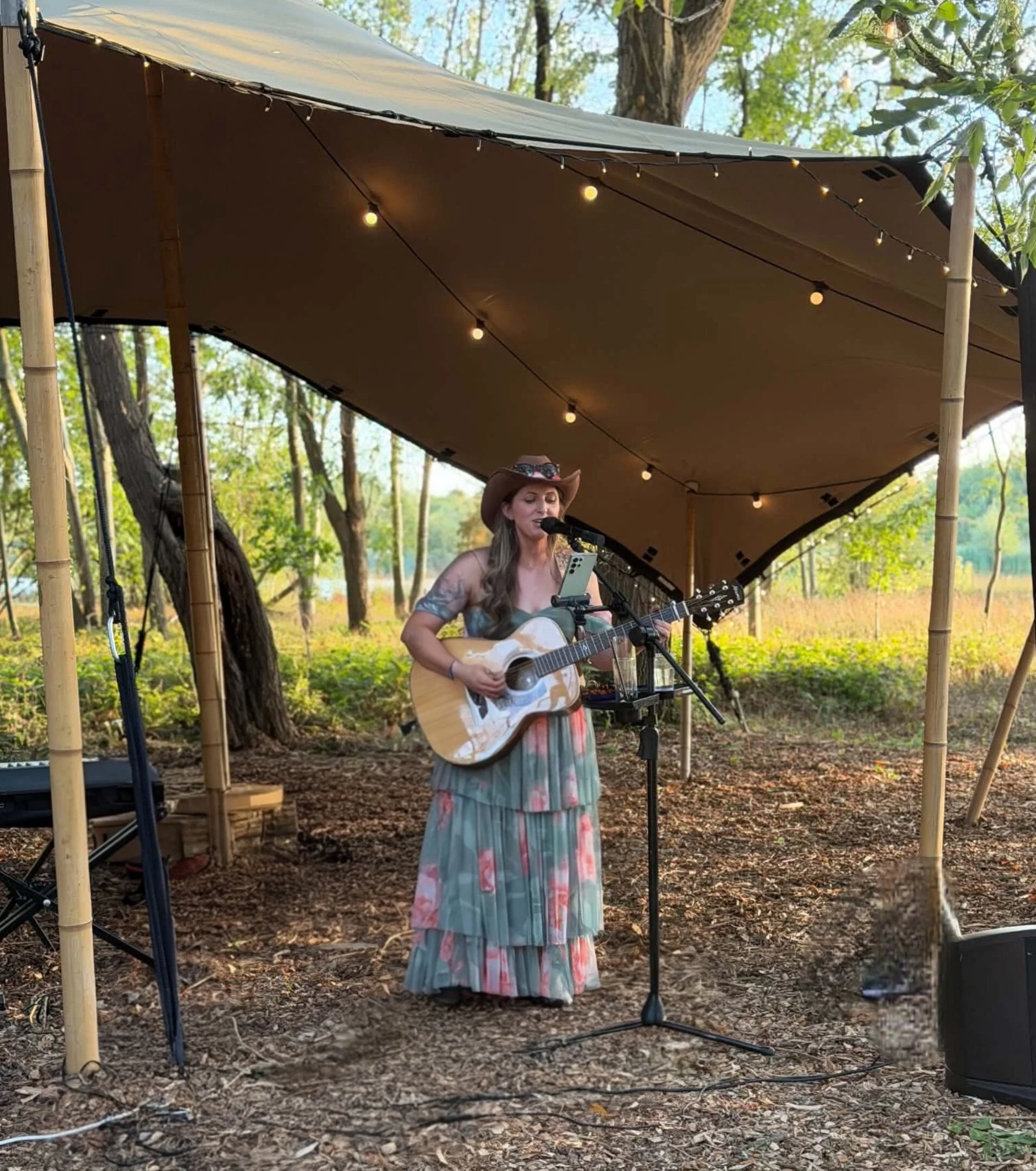 A woman wearing a cowboy hat and a tiered green dress is singing and playing an acoustic guitar outdoors under a canopy with string lights, surrounded by trees with green leaves.