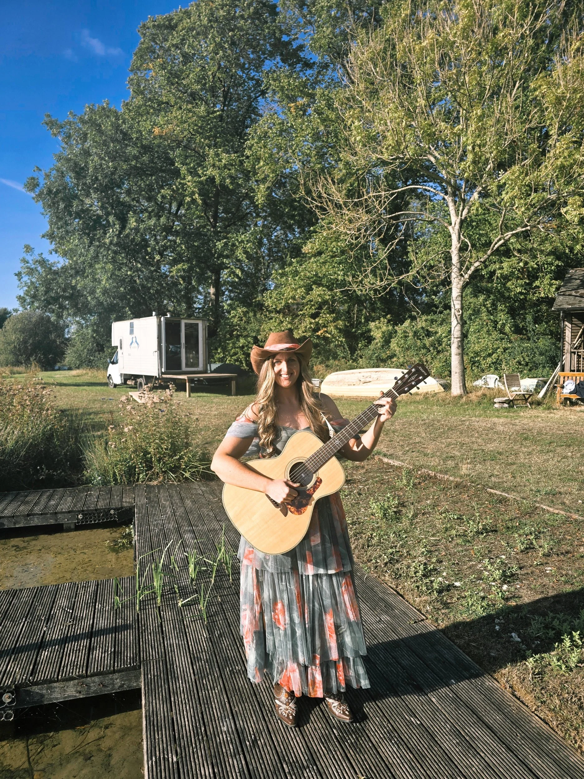 Woman with an acoustic guitar wearing a cowboy hat and boots, a green and orange floral tuille dress with long wavy brown hair smiling. Stood on a dock next to a lake with trees and cabins in the background.
