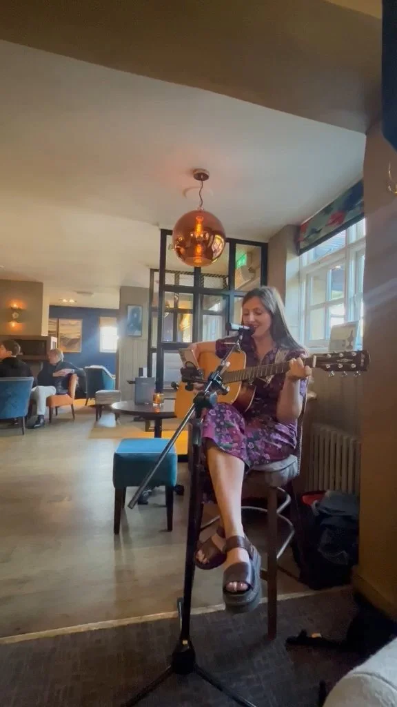 A woman singing and playing guitar at a pub, with a microphone in front of her, in a cozy, wood-paneled setting.