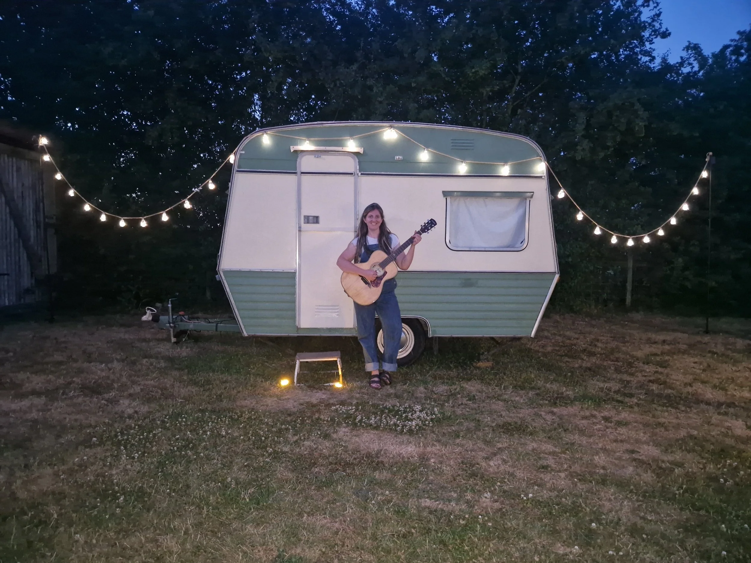 A young woman smiling and playing an acoustic guitar in front of a vintage camper trailer decorated with string lights at dusk or night.