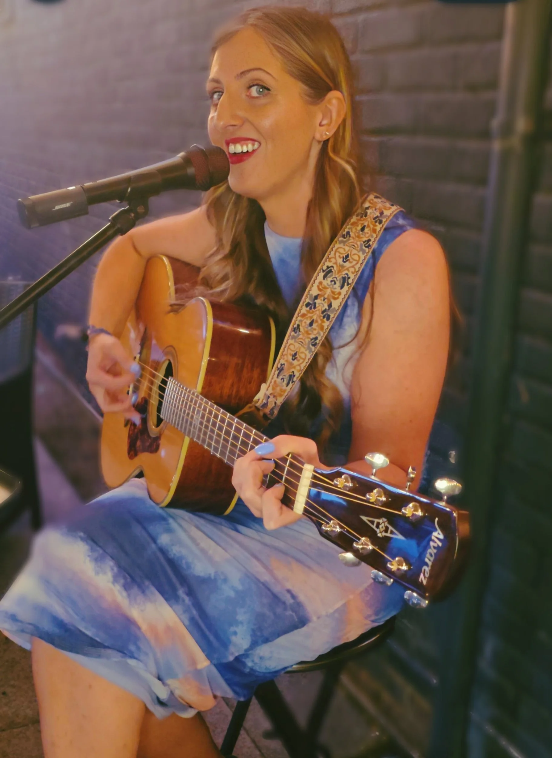 A woman playing an acoustic guitar and singing into a microphone, sitting against a brick wall.