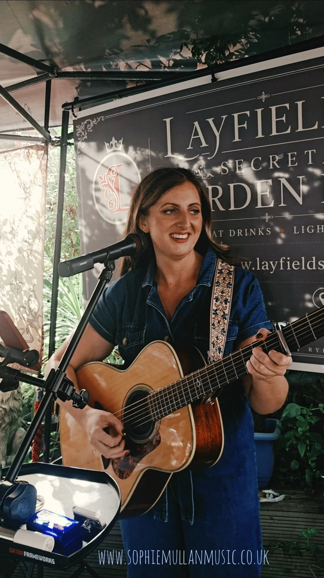 A woman playing an acoustic guitar and singing into a microphone at an outdoor event, with a black banner behind her that reads 'Layfield Secret Garden'