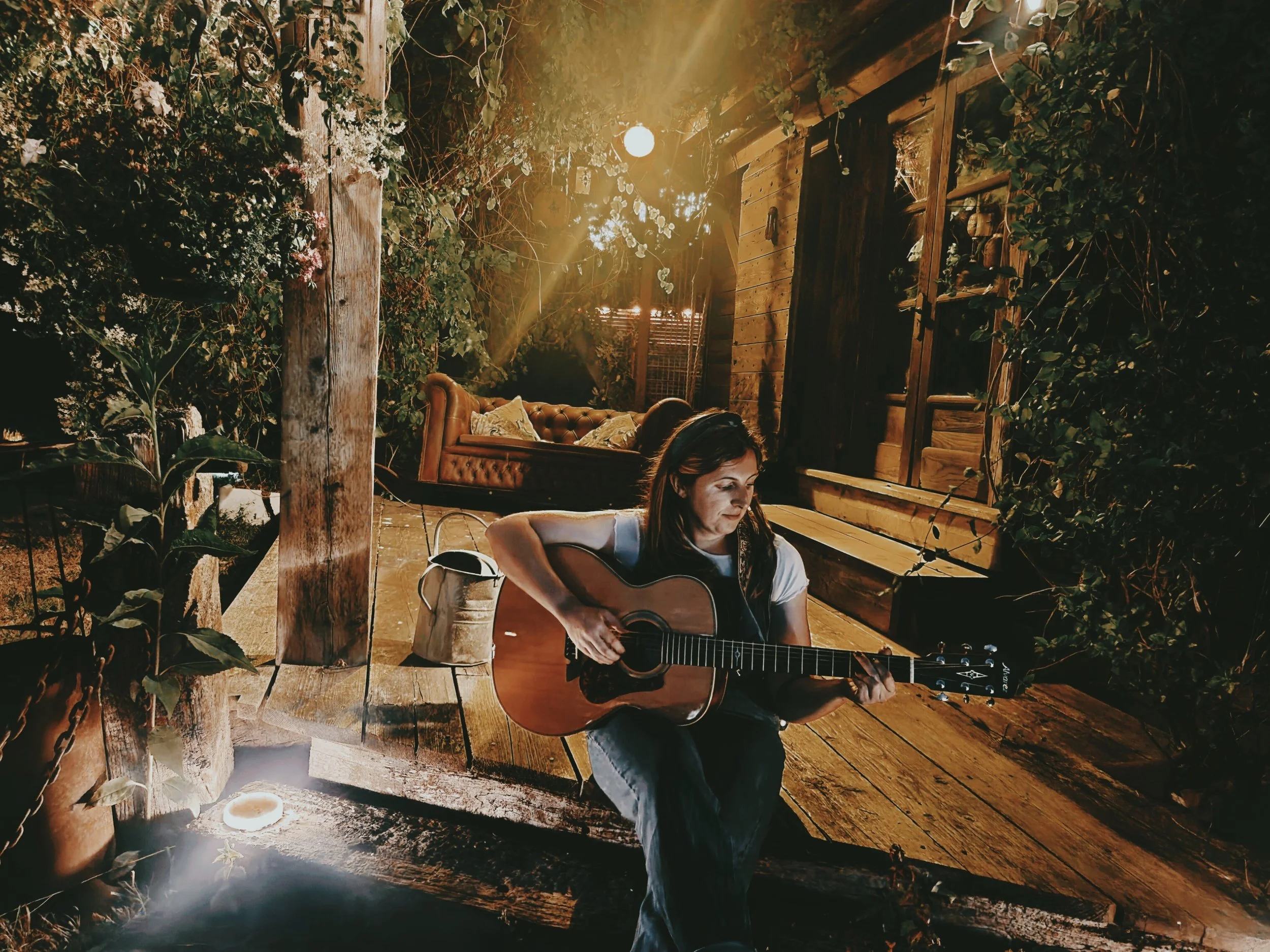 A woman with long dark hair playing an acoustic guitar, sitting on a cozy rustic deck covered by a canopy or green leaf foliage a swing sofa behind and a cabin to the right, and warm lighting.