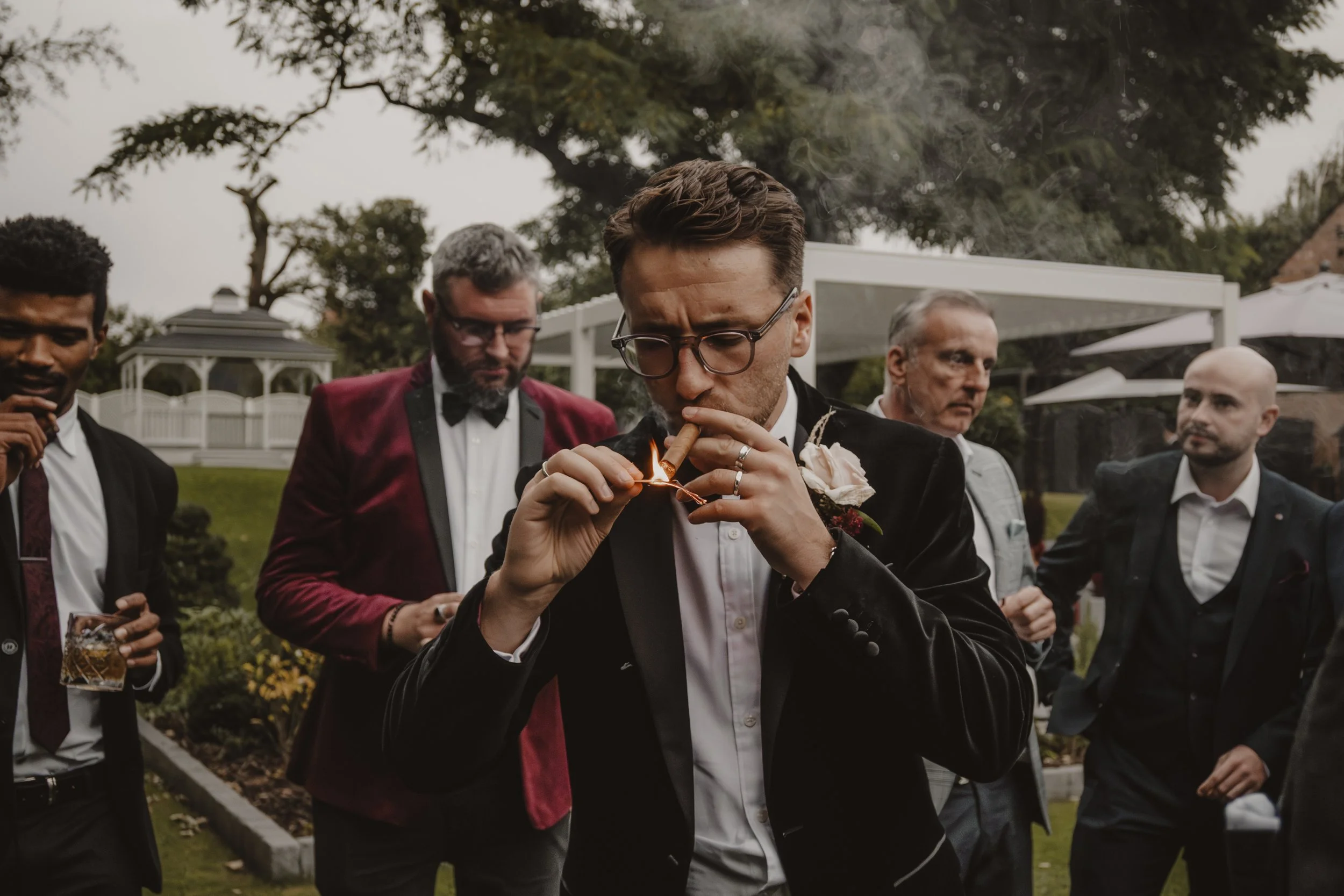 Groom in tuxedo lighting a cigar with a match at an outdoor gathering, surrounded by well-dressed men, in a garden setting.