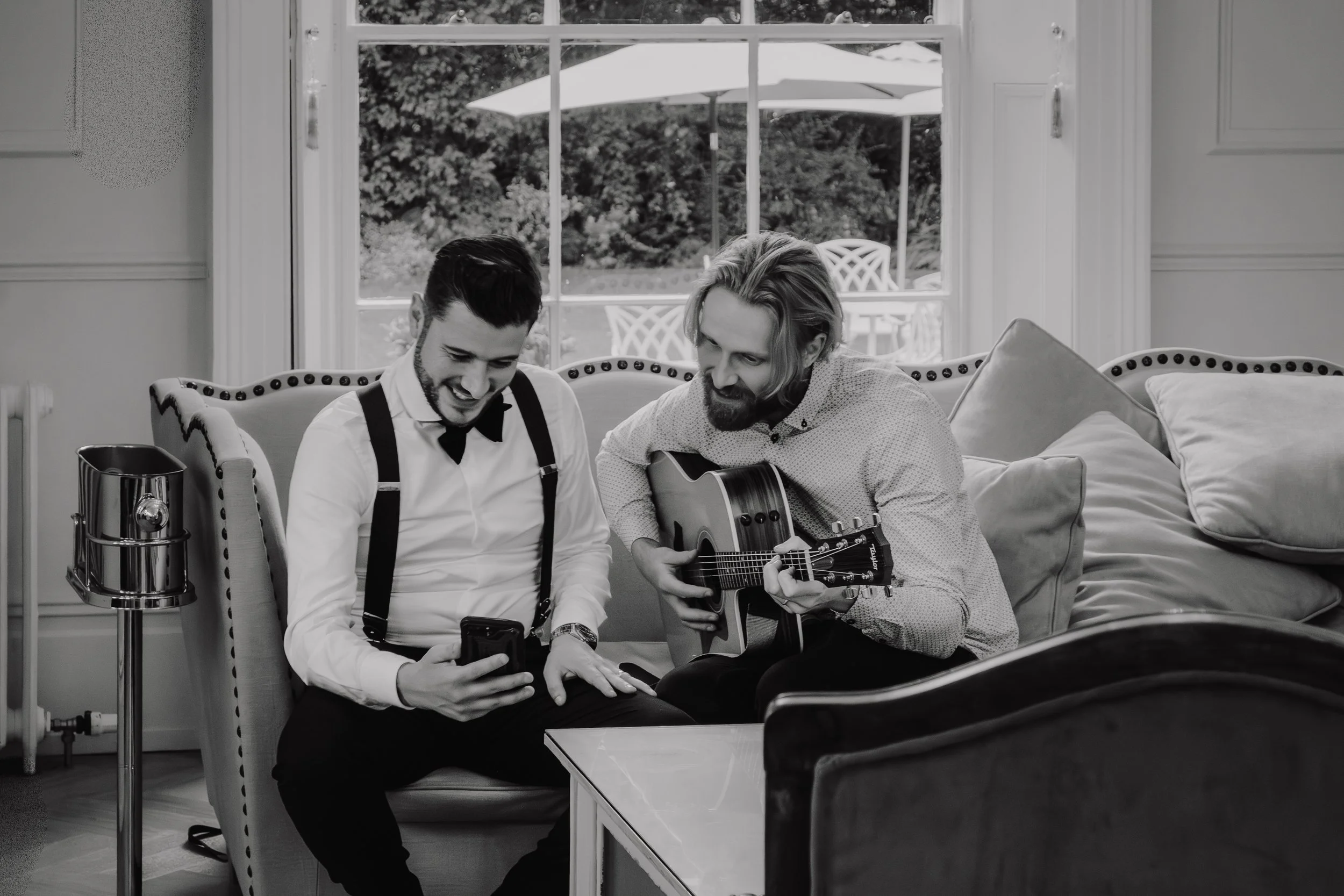 Two men sitting on a sofa in a living room, one is looking at a cell phone, the other is playing an acoustic guitar, with a large window and outdoor patio in the background.