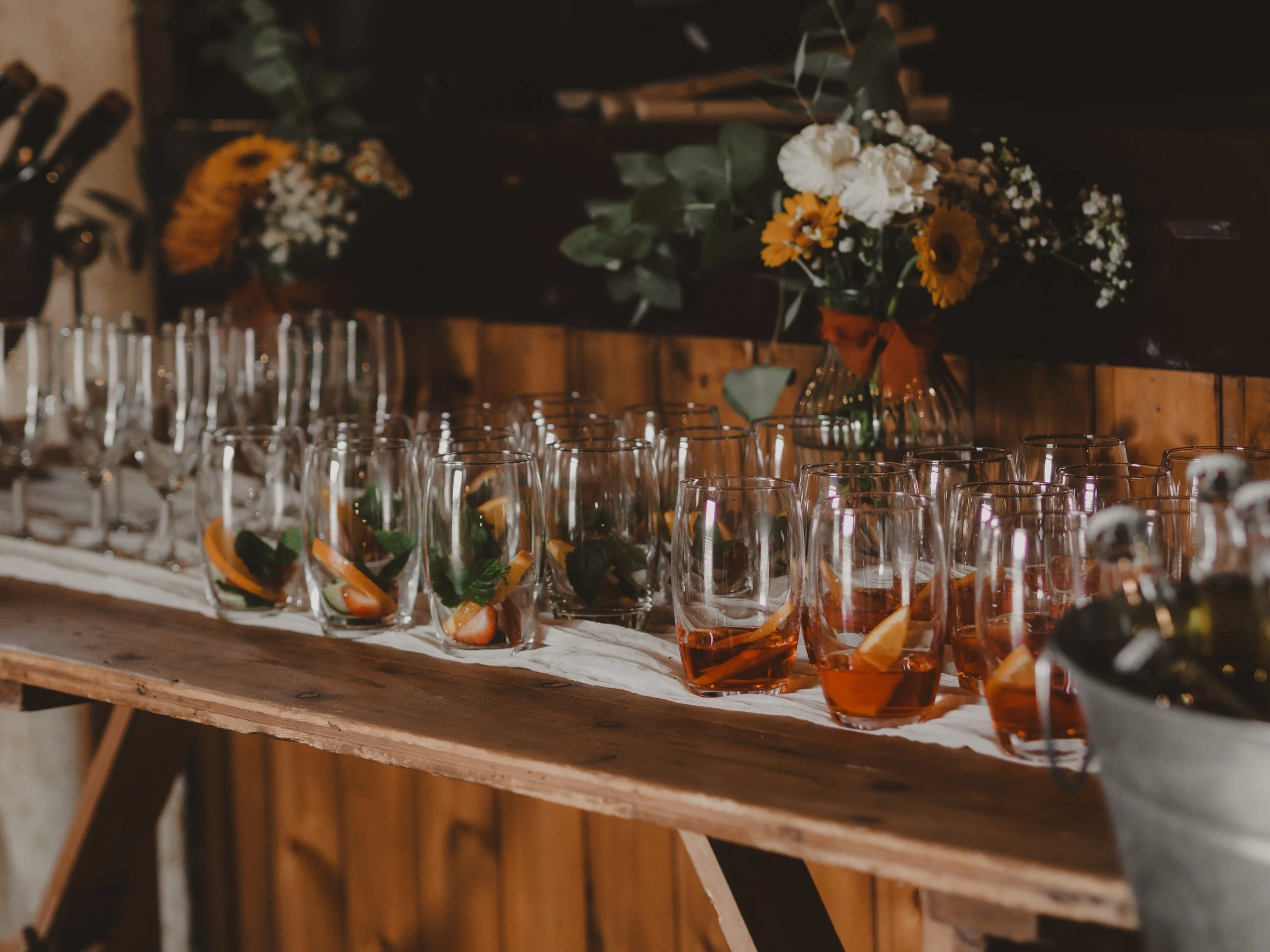 A rustic wooden table with glasses of an orange drink garnished with fruit slices and mint leaves, and a floral arrangement with white and yellow flowers in a vase in the background.