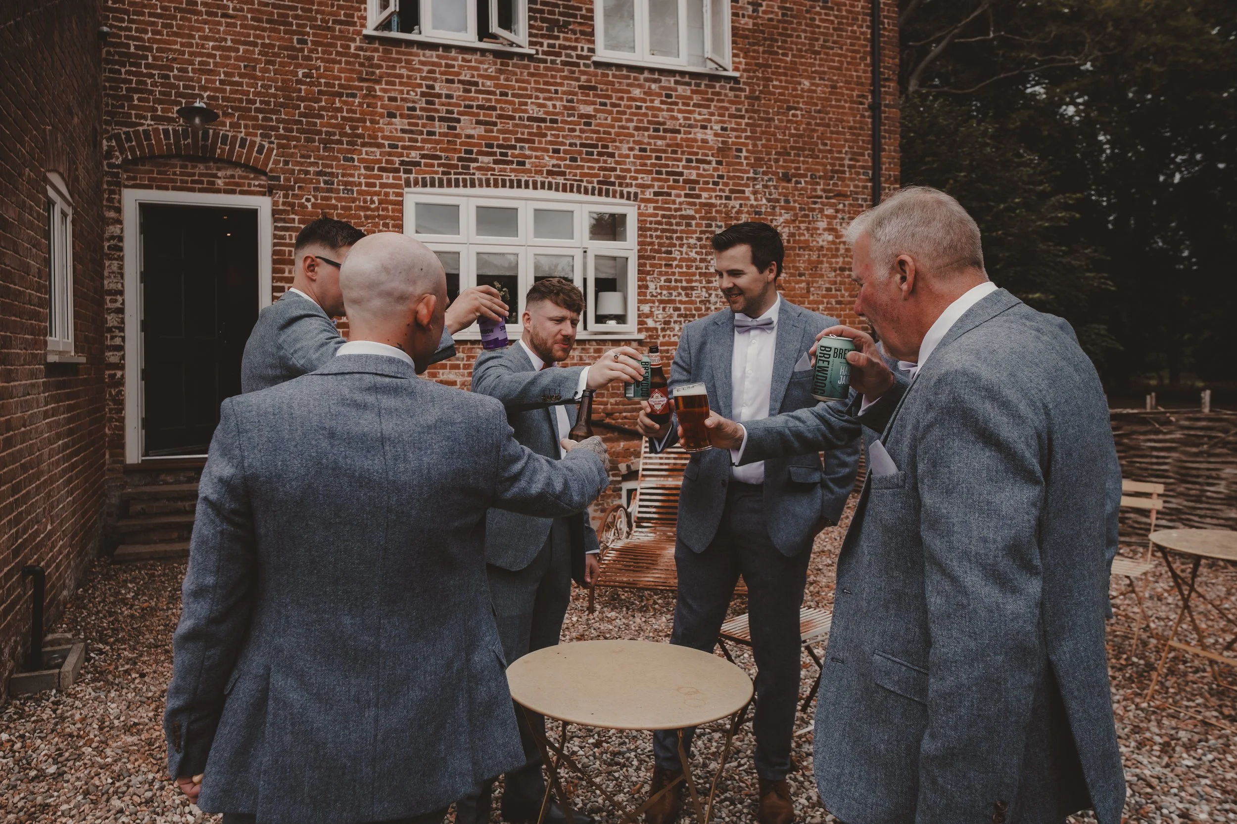 Candid photo of Groomsmen in grey suits having drinks pre wedding ceremony outdoors in a backyard with brick house in the background.