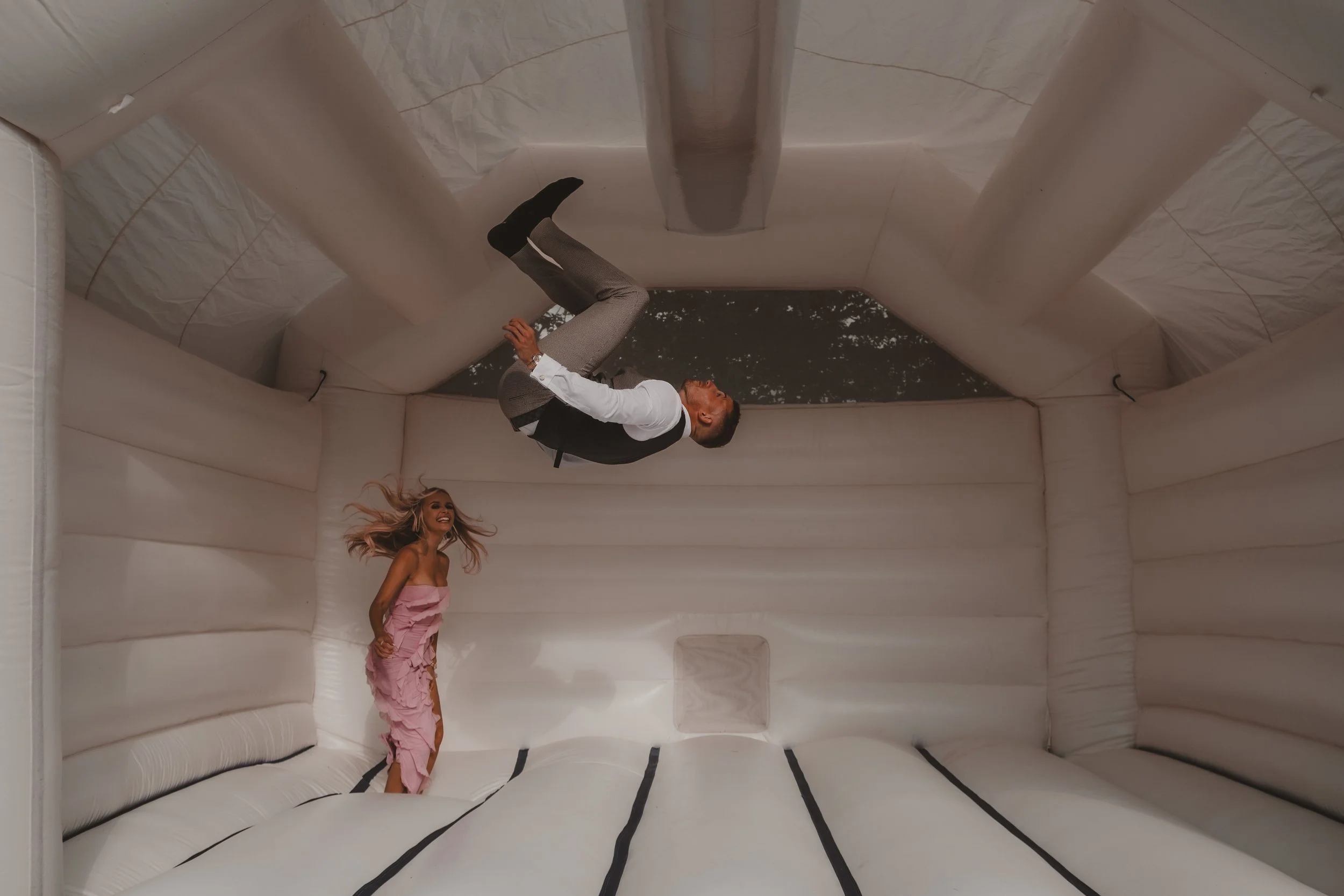 A man and a woman inside an inflatable bounce house. The man is upside down, wearing a gray suit and black vest, and the woman is standing and laughing, wearing a pink ruffled dress. The interior of the bounce house is white with black lines on the f
