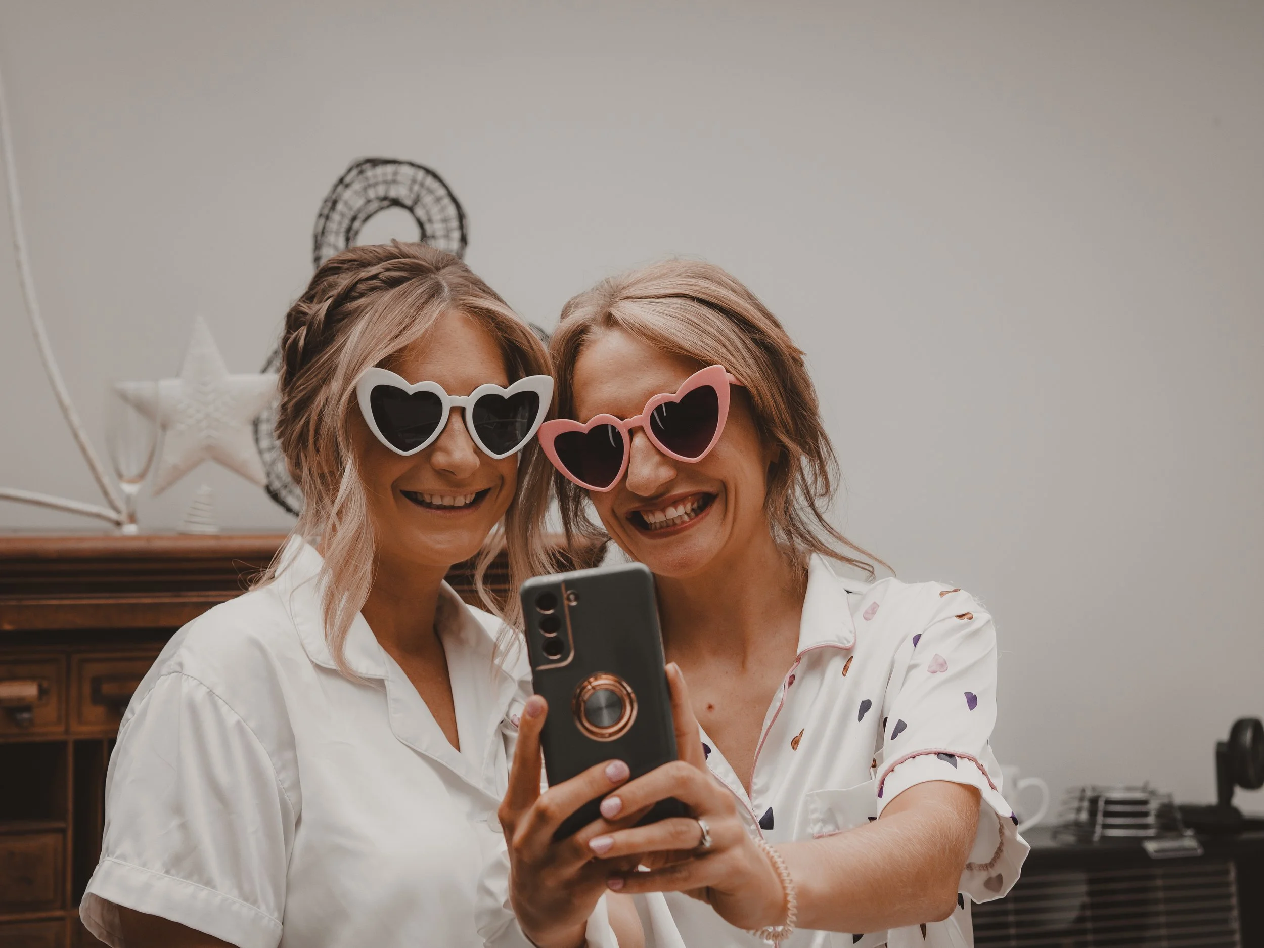 Two women with blonde hair wearing heart-shaped sunglasses taking a selfie together indoors.