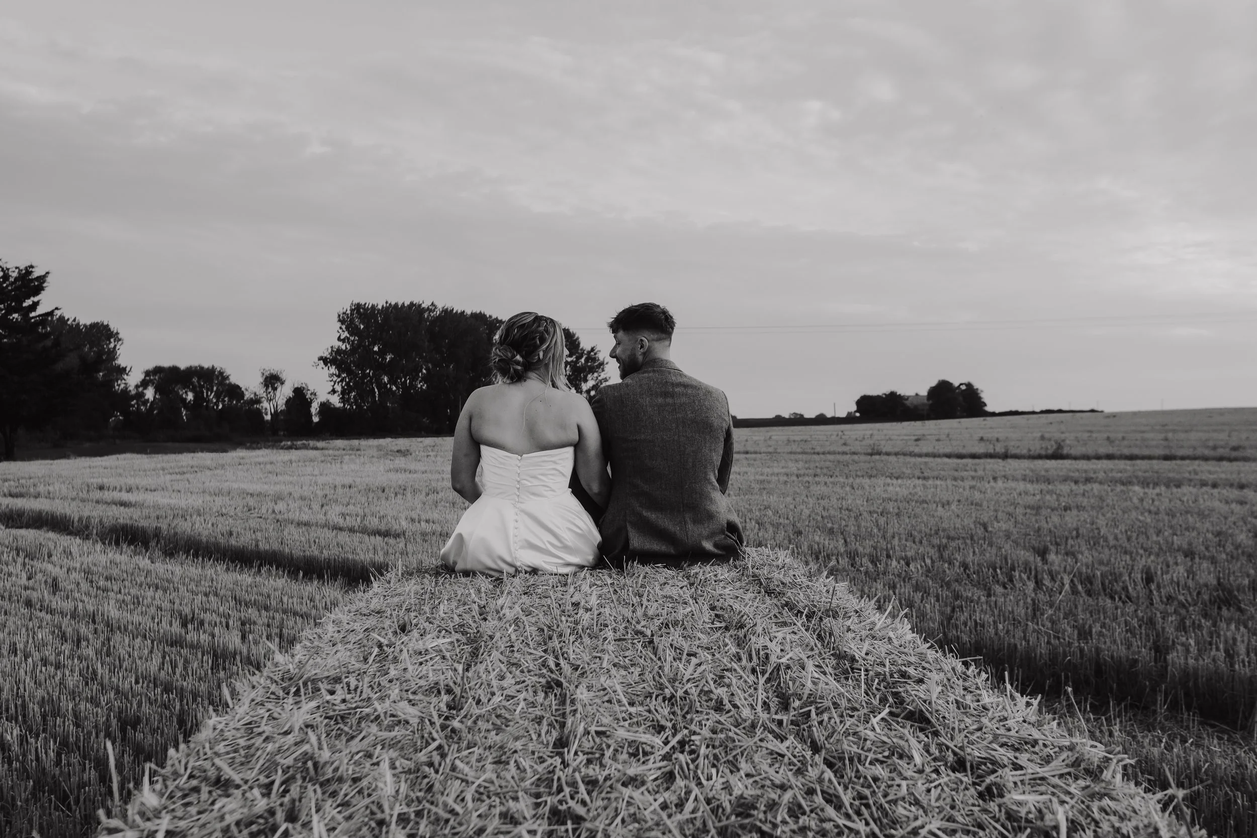 A black-and-white photo of a couple sitting together on a haystack in a field, facing away from the camera and looking at each other, with trees and a cloudy sky in the background.