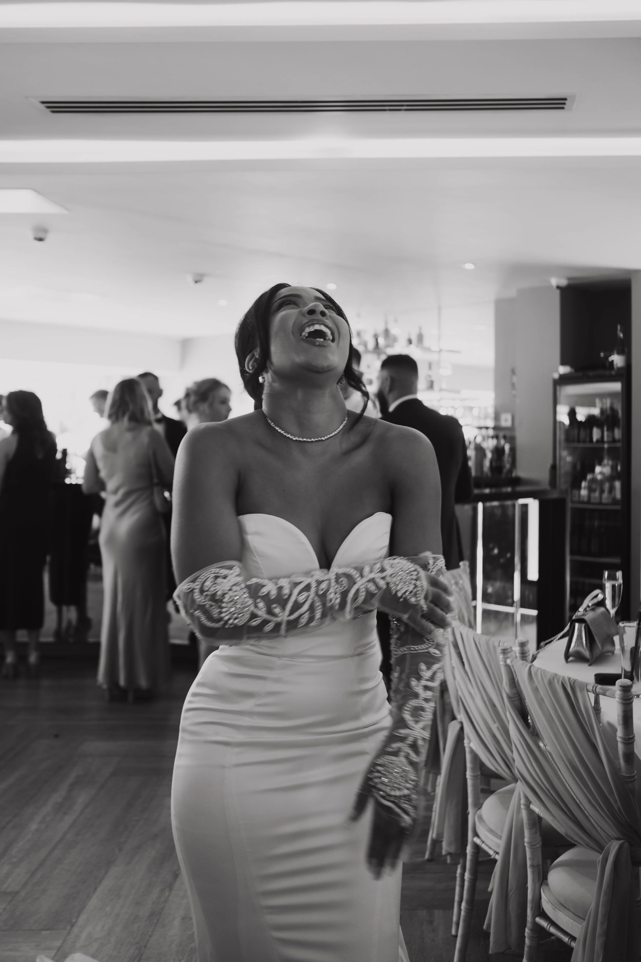 A woman in a strapless wedding dress with lace gloves laughing at a wedding reception, with guests and a bar in the background.