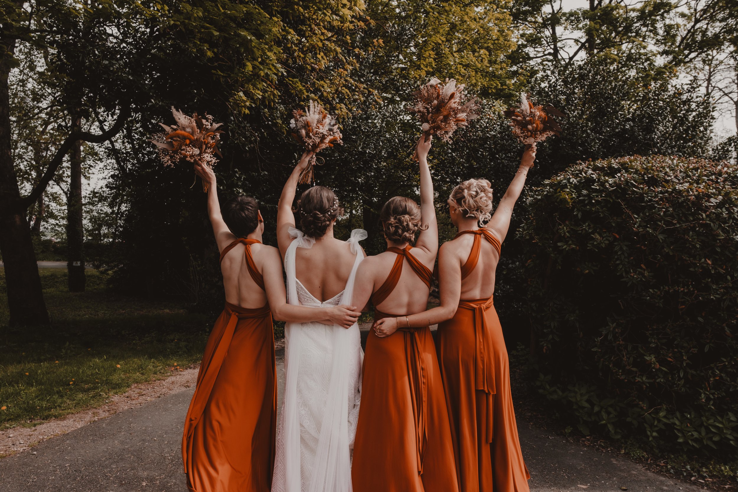 A bride and three bridesmaids standing outdoors with their backs to the camera, holding bouquets, with trees in the background.