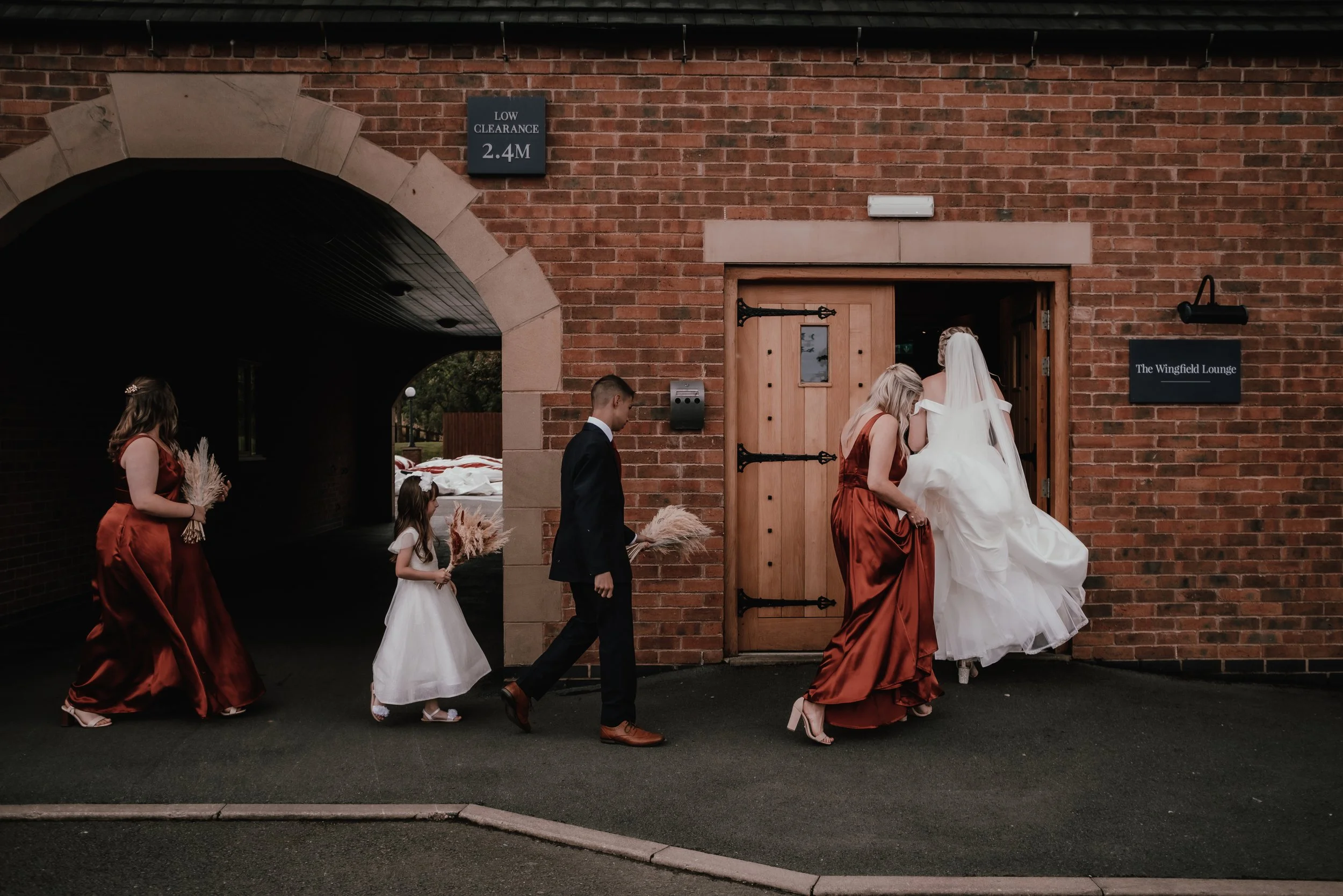 A wedding procession leaving a building called The Wingfield Lounge. The bride in a white gown is walking up steps with two women in bronze-colored dresses, a young girl in a white dress with a bow, and a man in a dark suit carrying a bouquet. They a