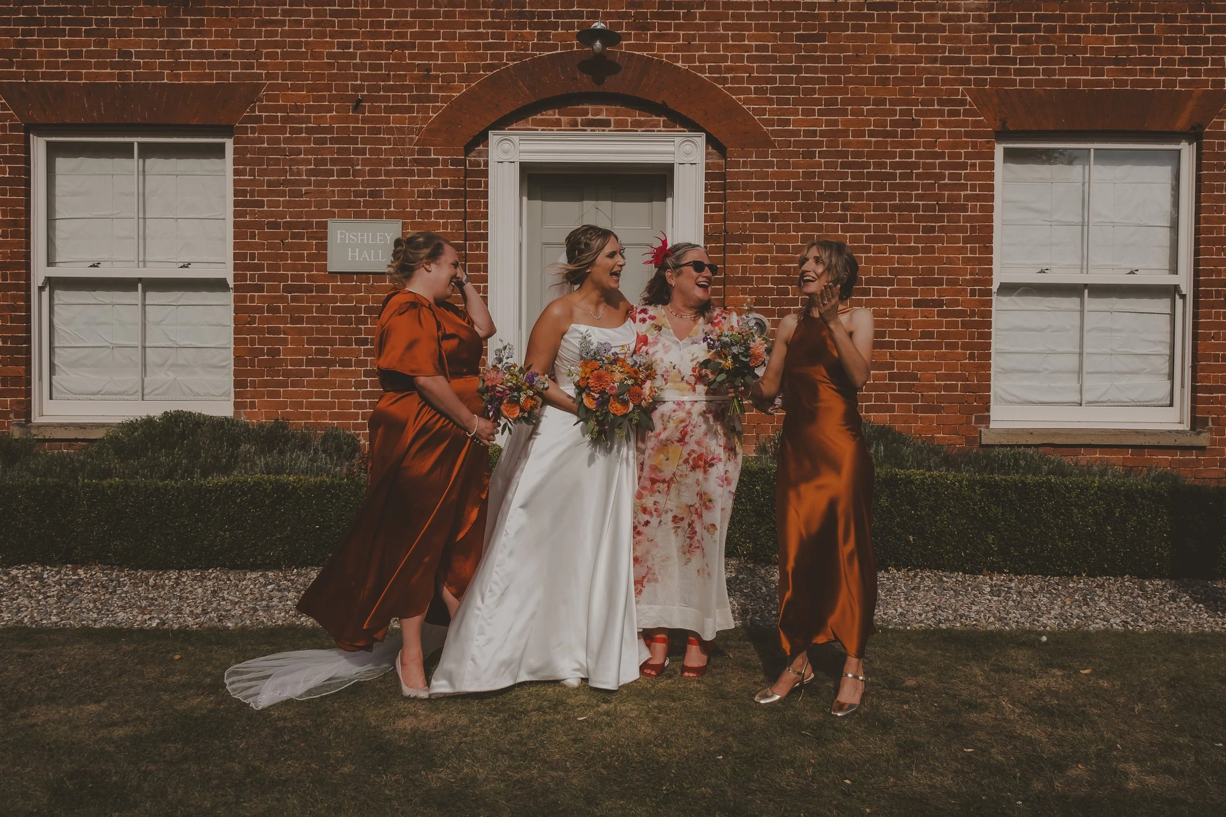 Bride and bridesmaids holding a bouquet, are standing outside a brick building with a sign that reads "Fishley Hall," laughing and chatting.
