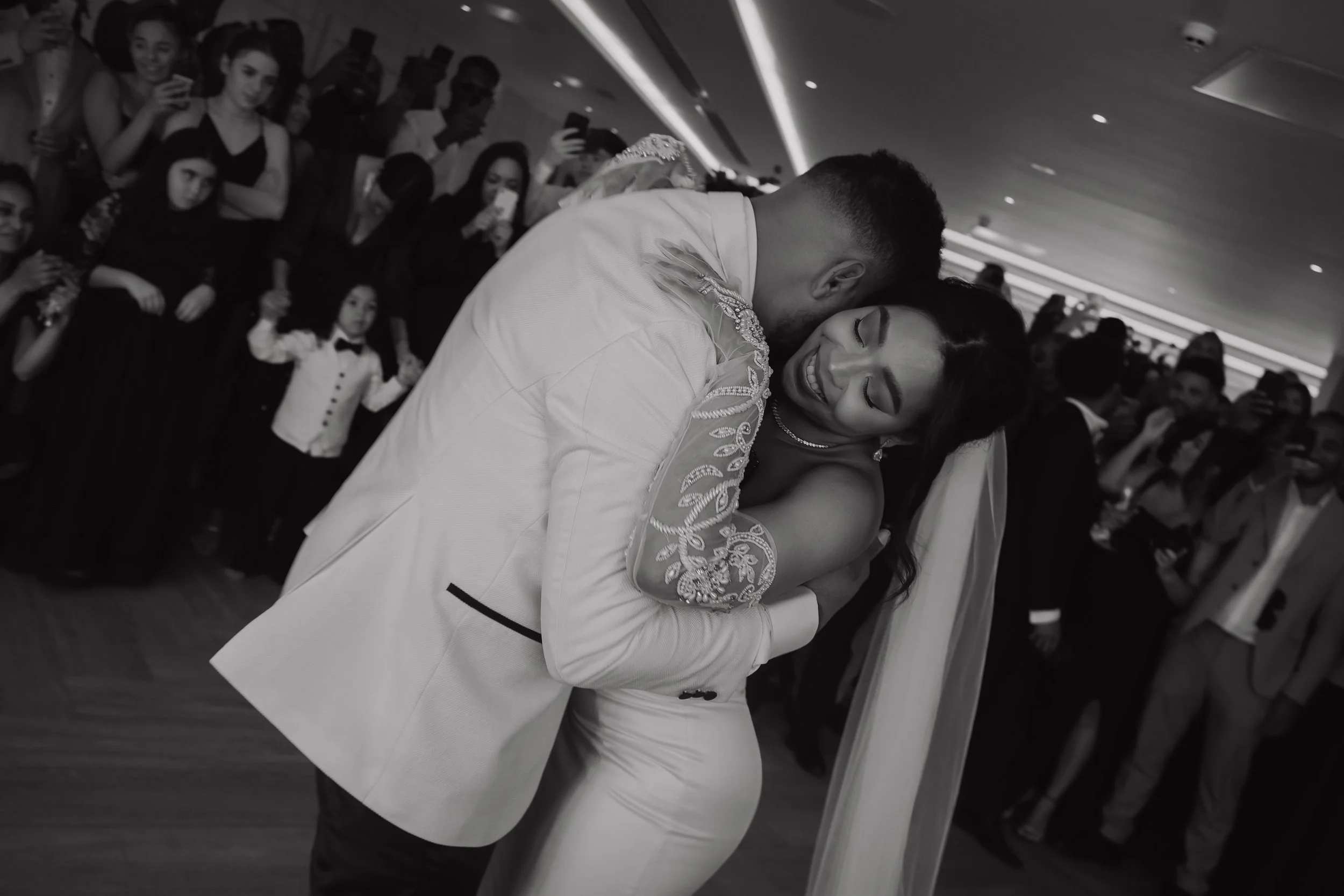 A couple dancing closely in a wedding reception, surrounded by guests taking photos and watching, in a stylish indoor venue.