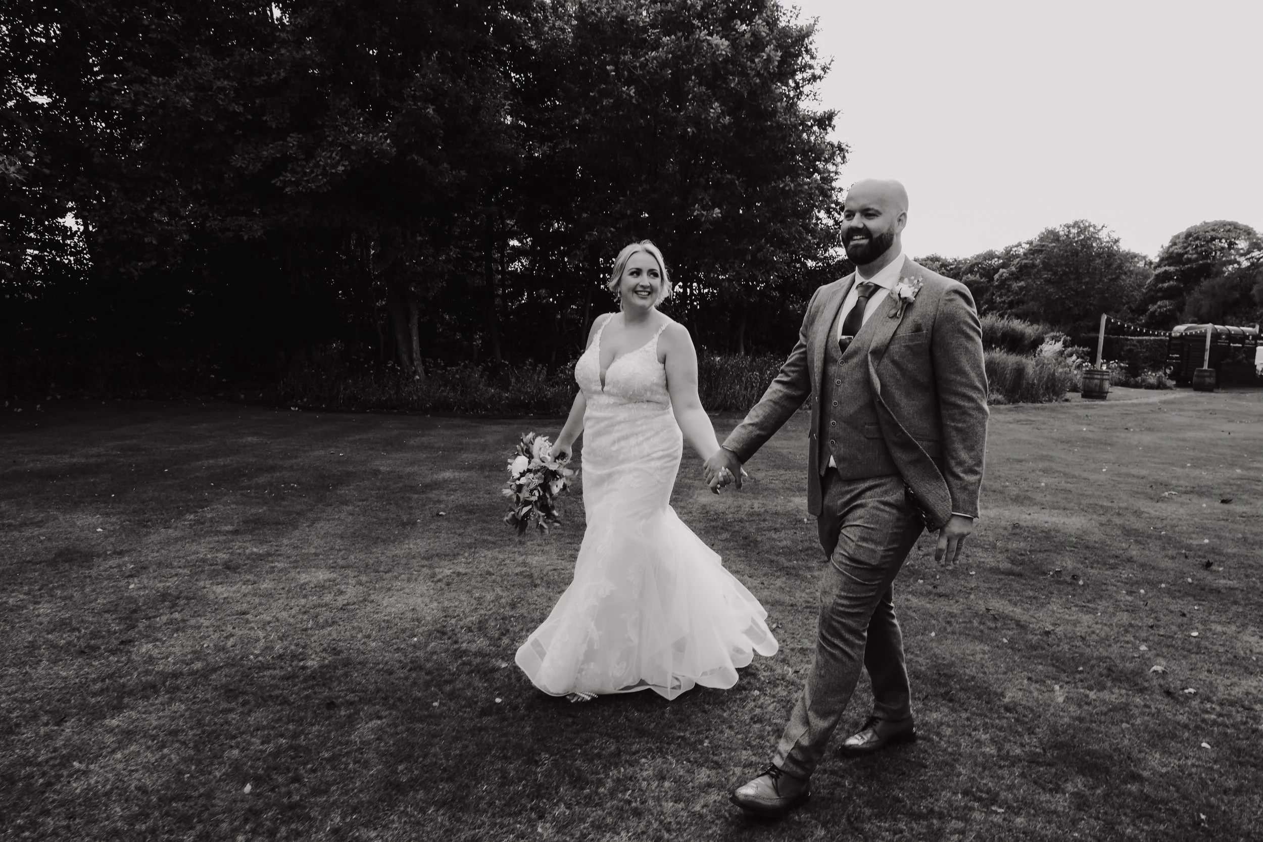 A black and white photo of a bride and groom holding hands and walking on a grassy field. The bride is wearing a lace wedding gown and holding a bouquet, while the groom is dressed in a suit with a tie and boutonniere. They are smiling and looking at