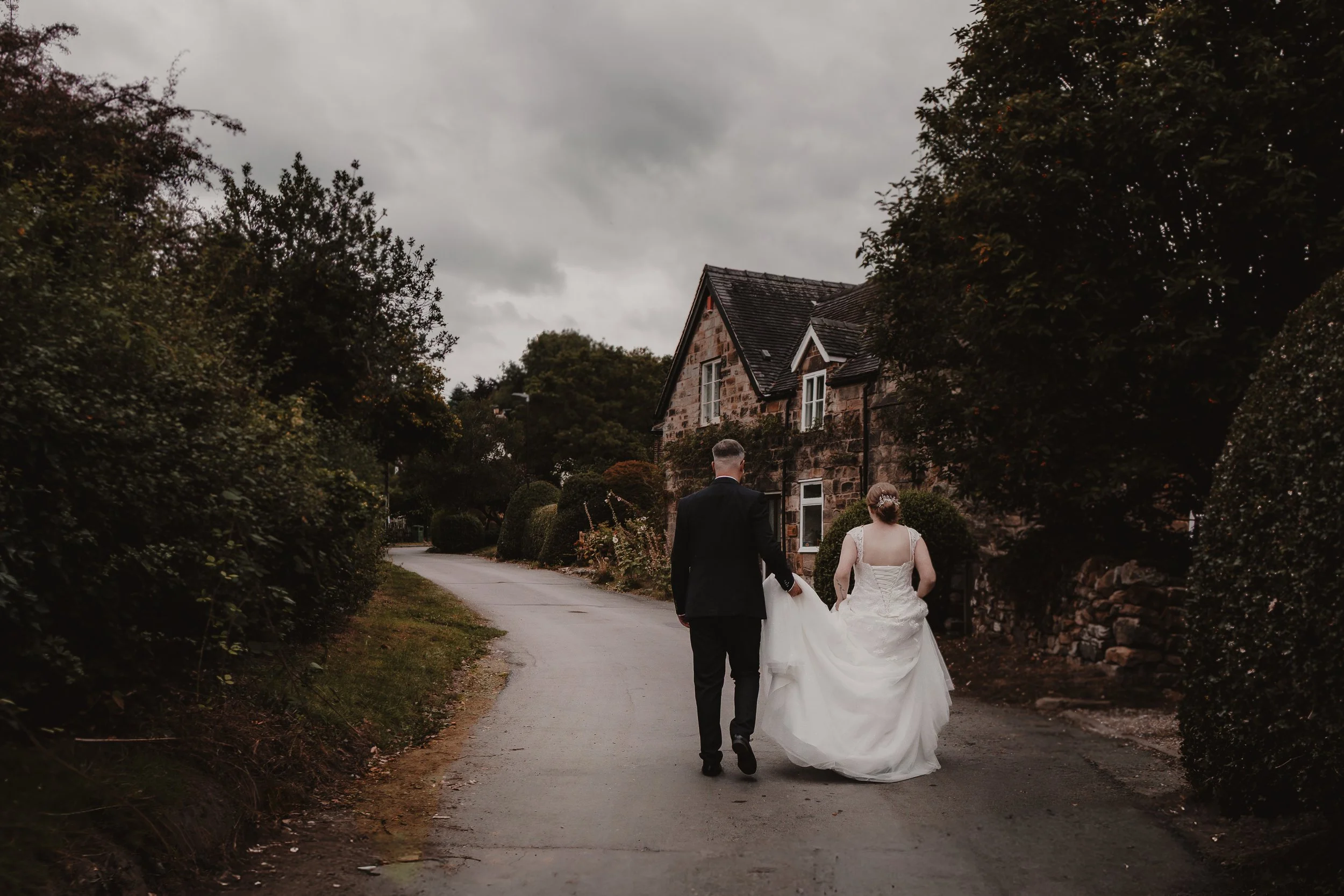 A bride and groom walking on a winding country road, surrounded by greenery and trees, with a stone house in the background under a cloudy sky.