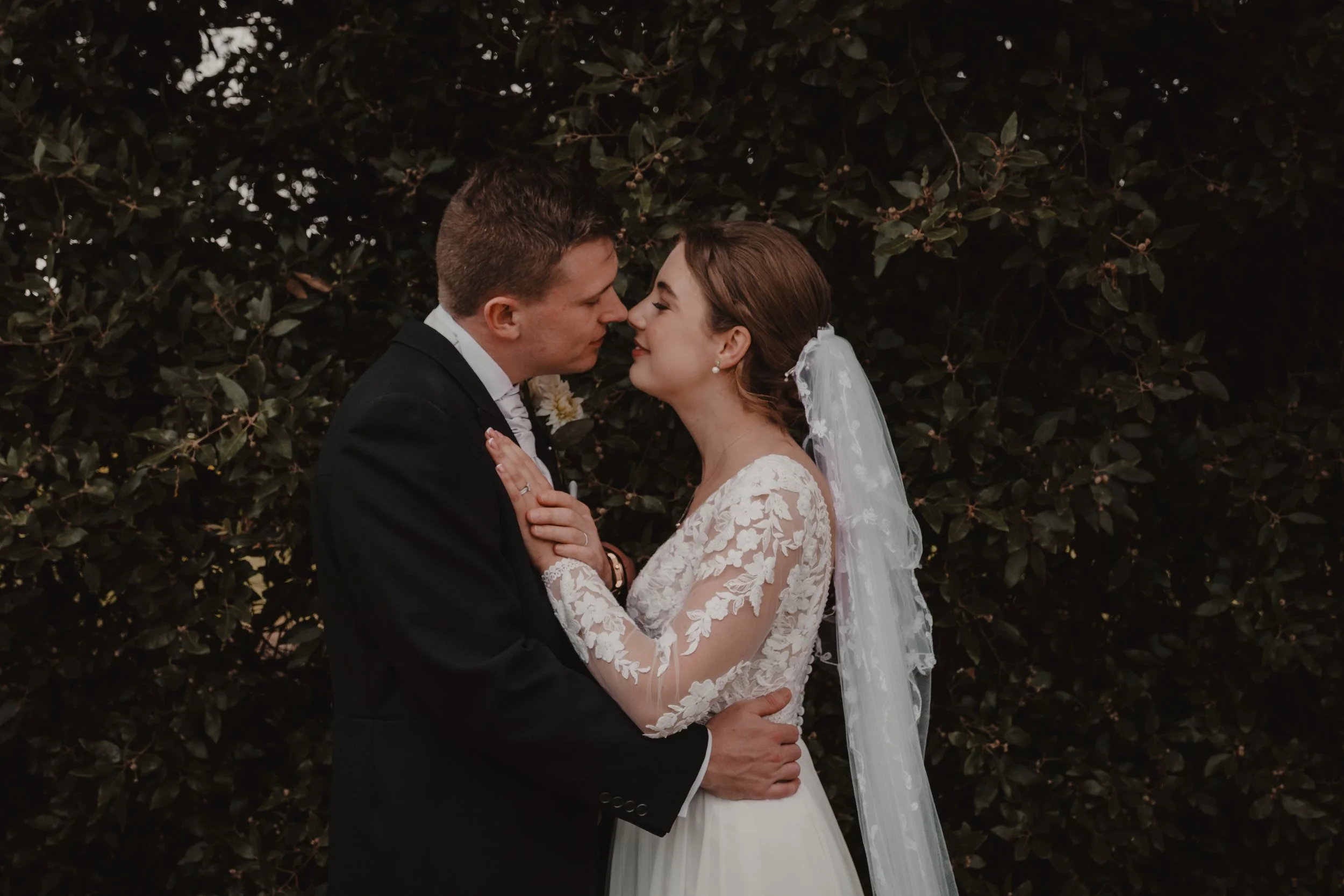 A bride and groom standing close together outdoors, face-to-face, with a dark leafy background, on their wedding day.