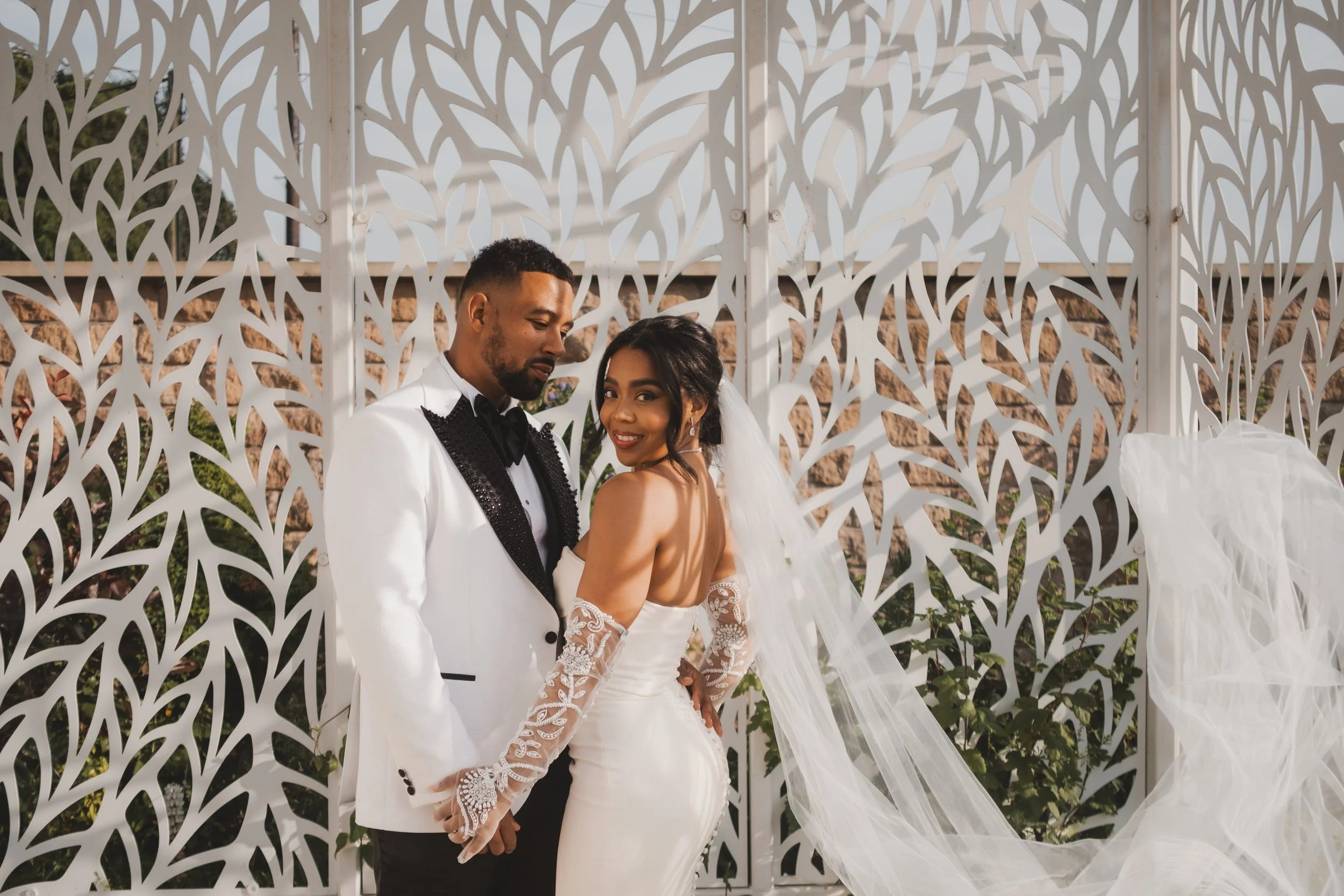 A bride and groom posing together at their wedding in front of a decorative white lattice backdrop with greenery.