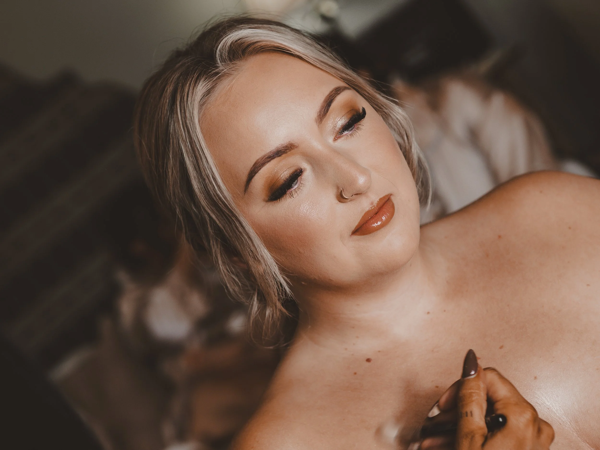 A woman with blonde hair getting her makeup done, looking serene, showing her bare shoulders and chest, with a hand gently touching her collarbone.