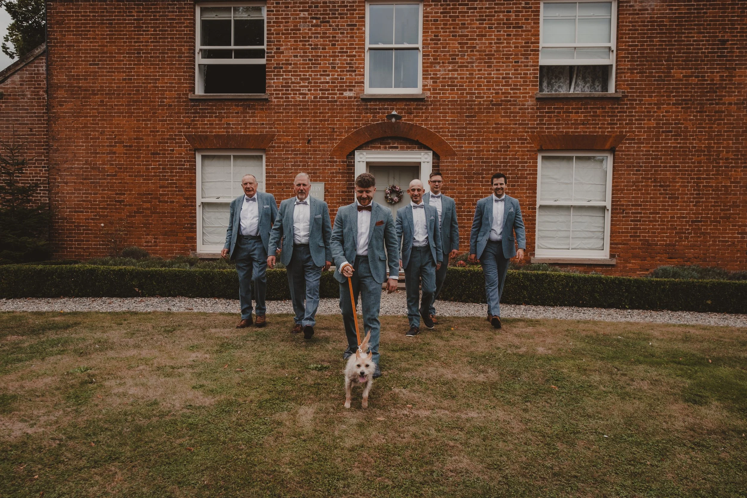 Six men dressed in matching blue suits and bow ties are walking on a lawn in front of a brick building. One man in the front is holding a leash attached to a small white dog. The men are smiling and appear to be happy.