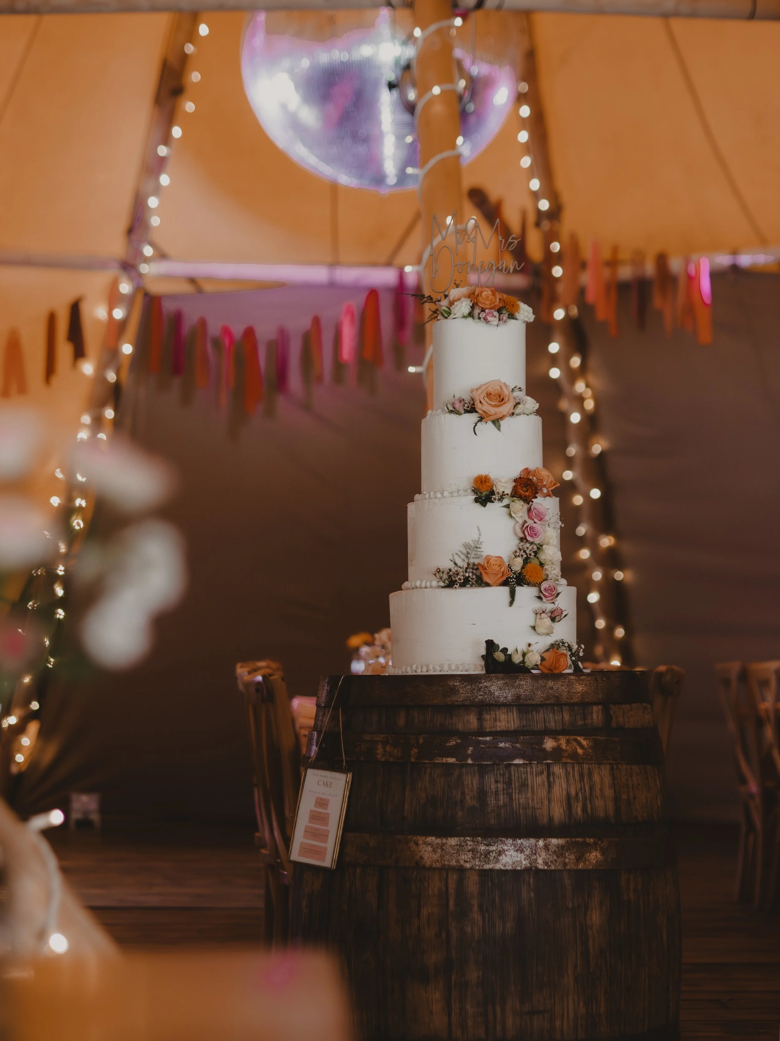 A four-tiered white wedding cake decorated with pink and orange flowers, placed on a wooden barrel under string lights at Whatton House Tipi in Loughborough.