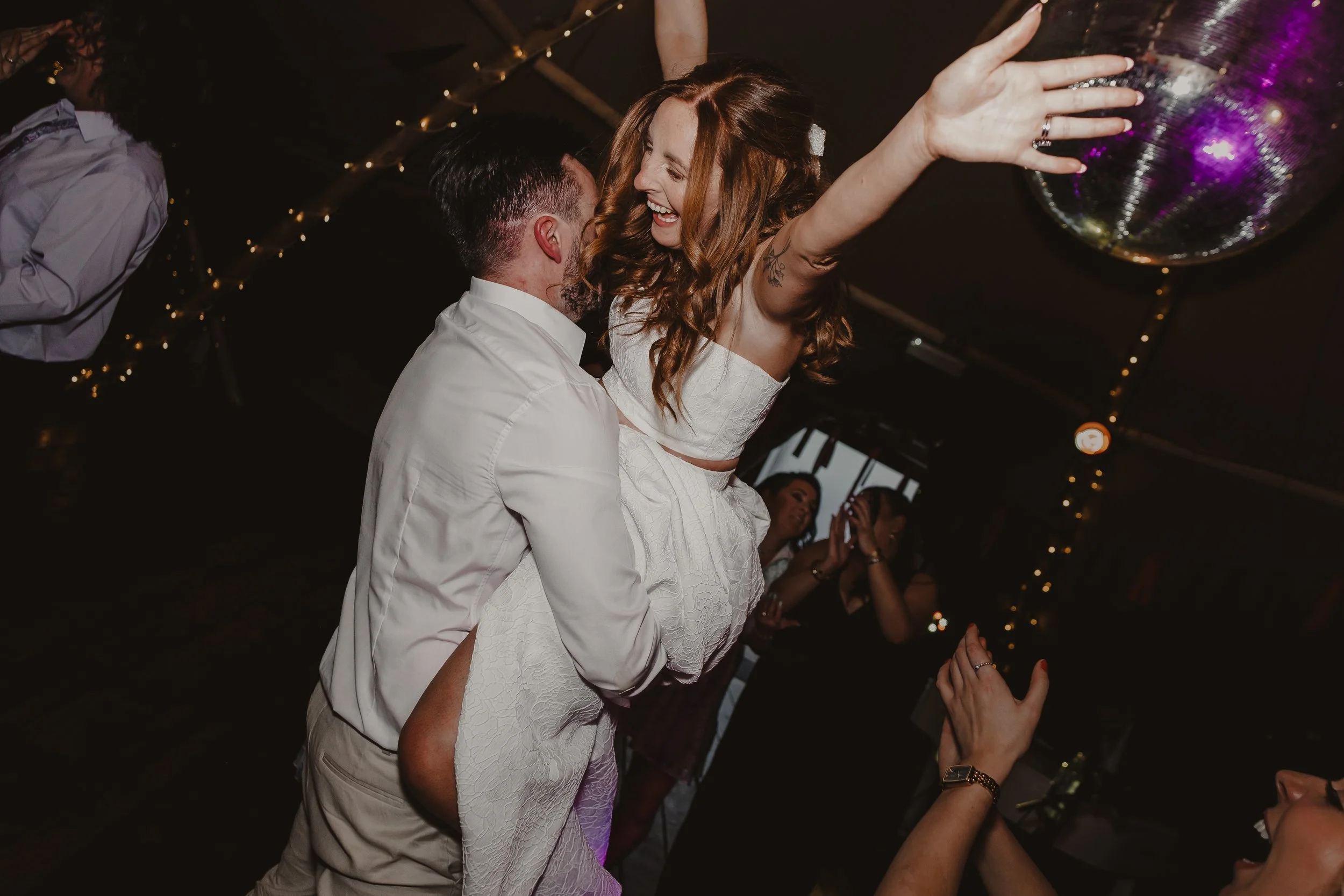 Bride and groom dancing, with the woman being lifted into the air, smiling and raising her arms. The venue is decorated with string lights and a disco ball.