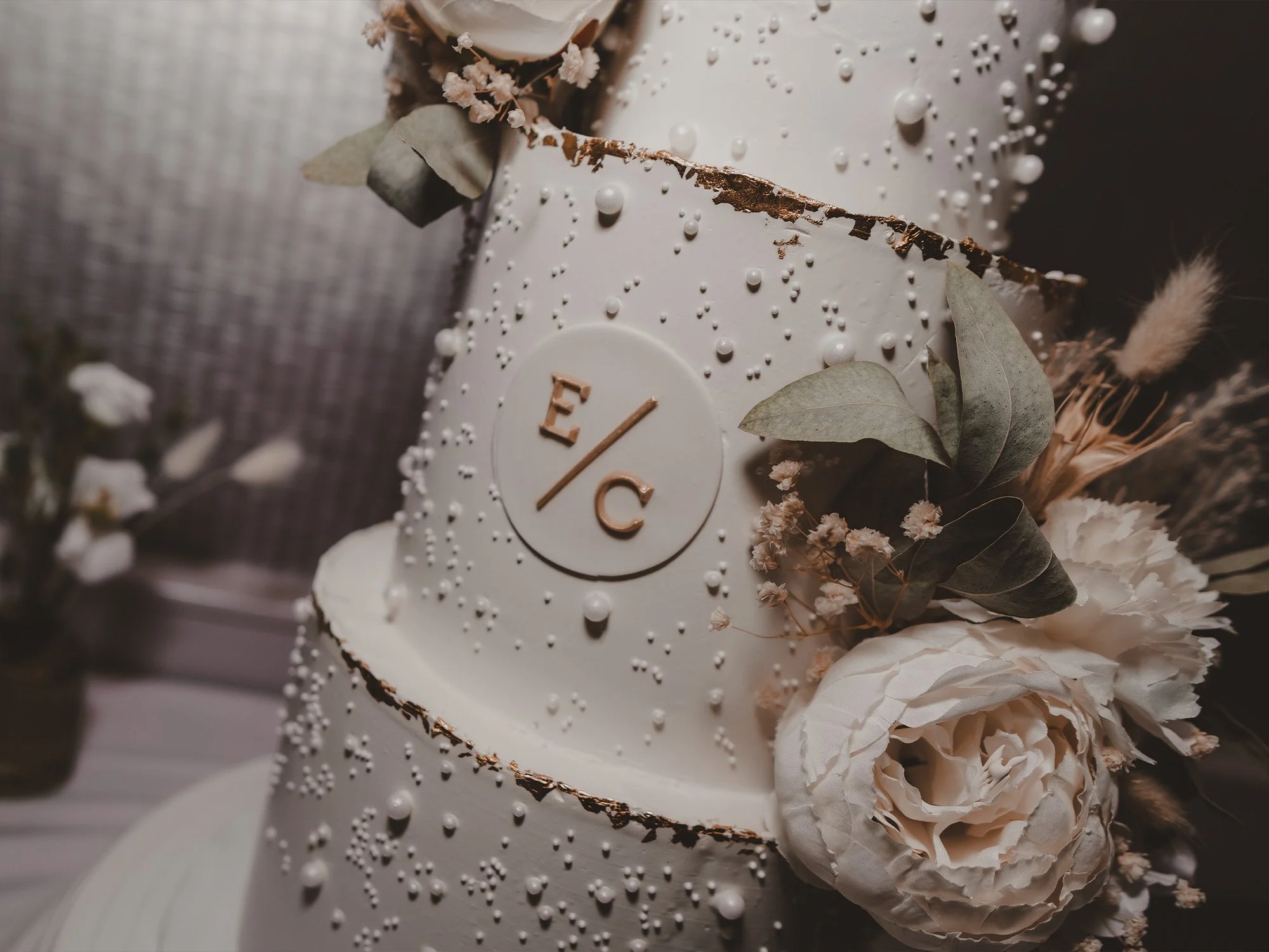 Close-up of a wedding cake decorated with white icing, small white pearl-like decorations, and gold accents, surrounded by white flowers and green leaves.