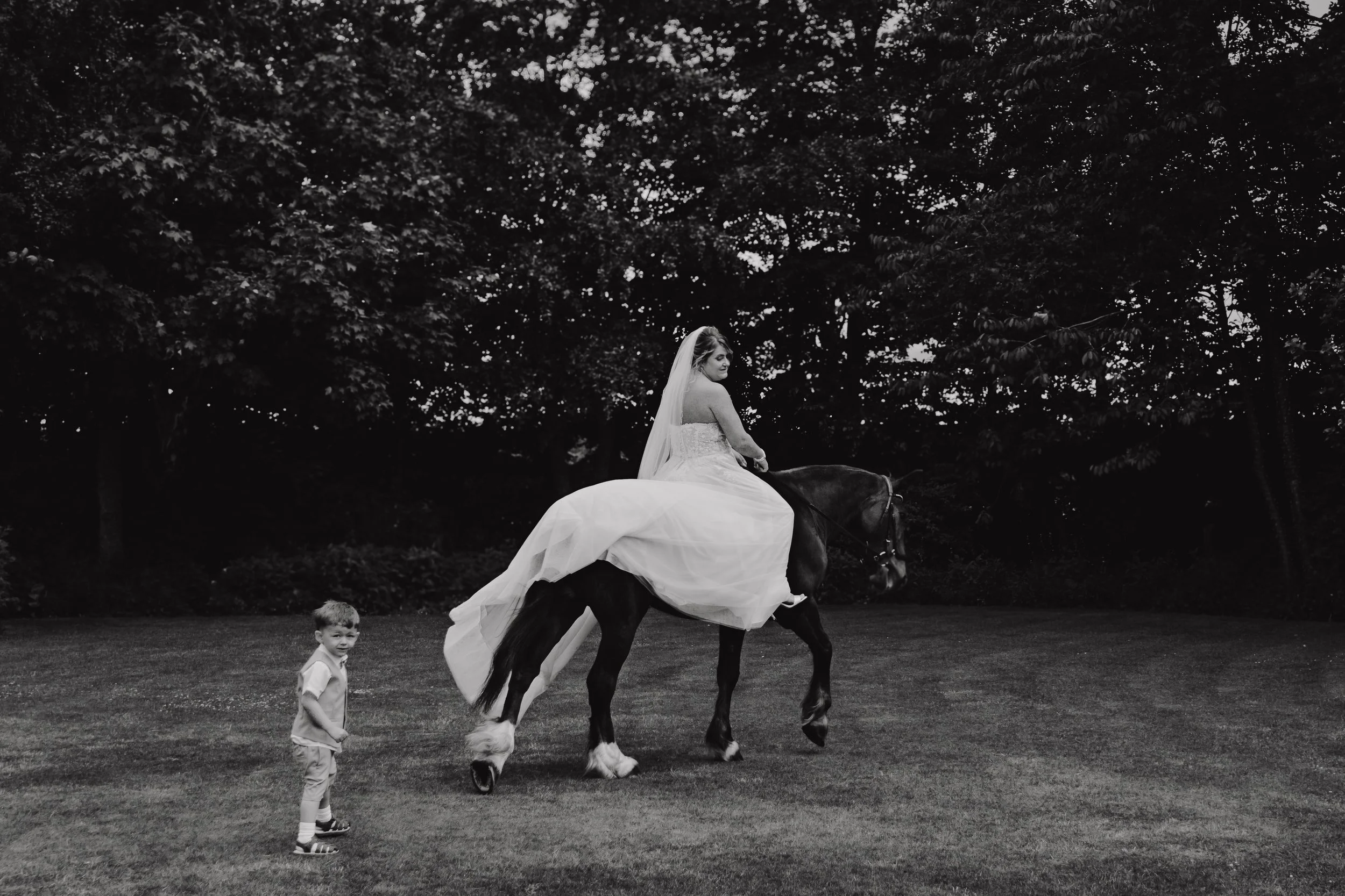 A black and white photograph of the bride in her wedding dress riding a horse in a park with trees in the background, while a young boy stands on the grass nearby looking at the camera.