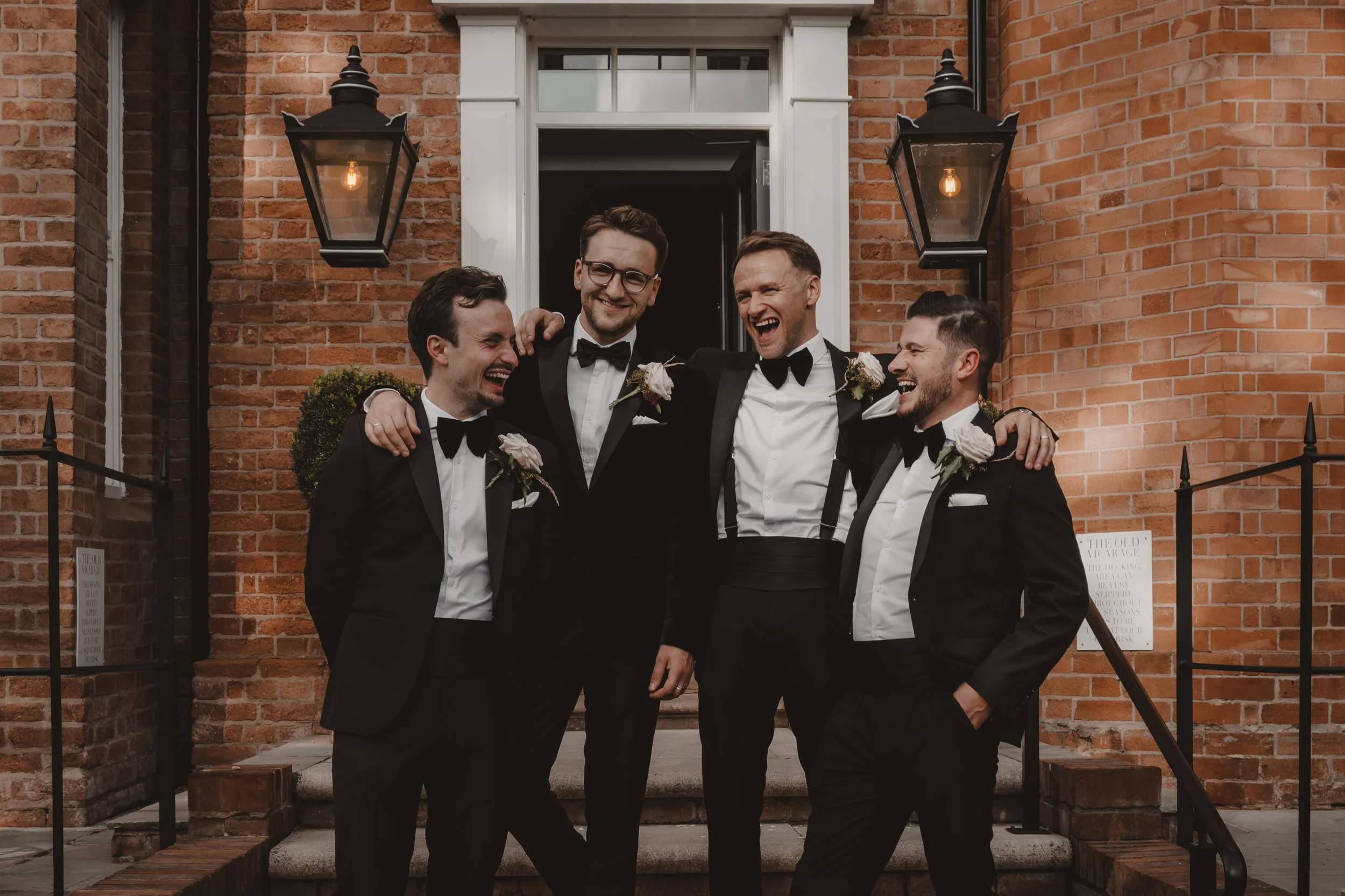 Groom and three groomsmen in tuxedos with boutonnières, smiling and laughing together on the steps in front of The Vicarage Boutique.