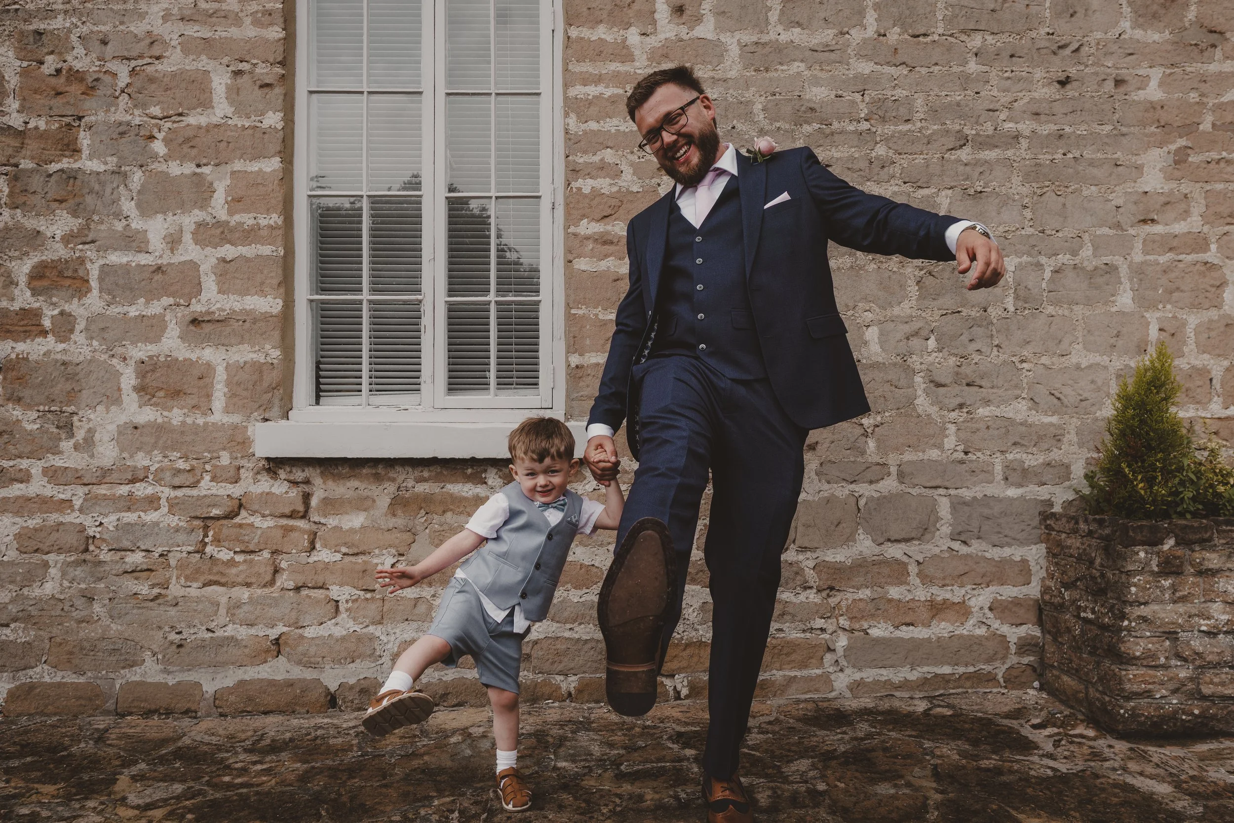 Groom in a navy suit and his young son in a light gray vest and shorts are playing together outside near a brick wall. The man is lifting his foot in a playful manner, and the boy is holding his hand, smiling and balancing on one leg.