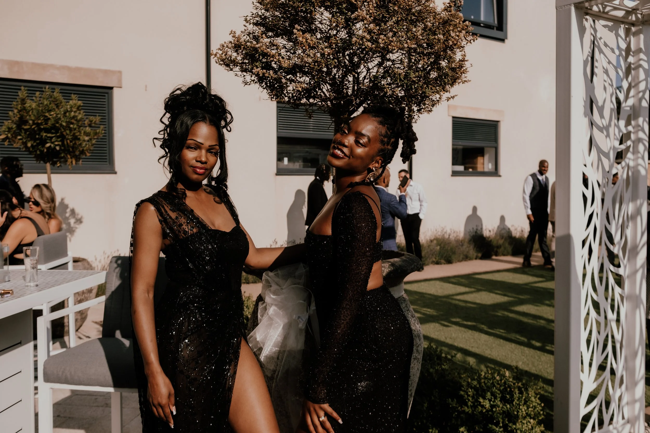Two women in black sparkly dresses smiling at a social gathering outdoors on a sunny day, with other guests in the background.