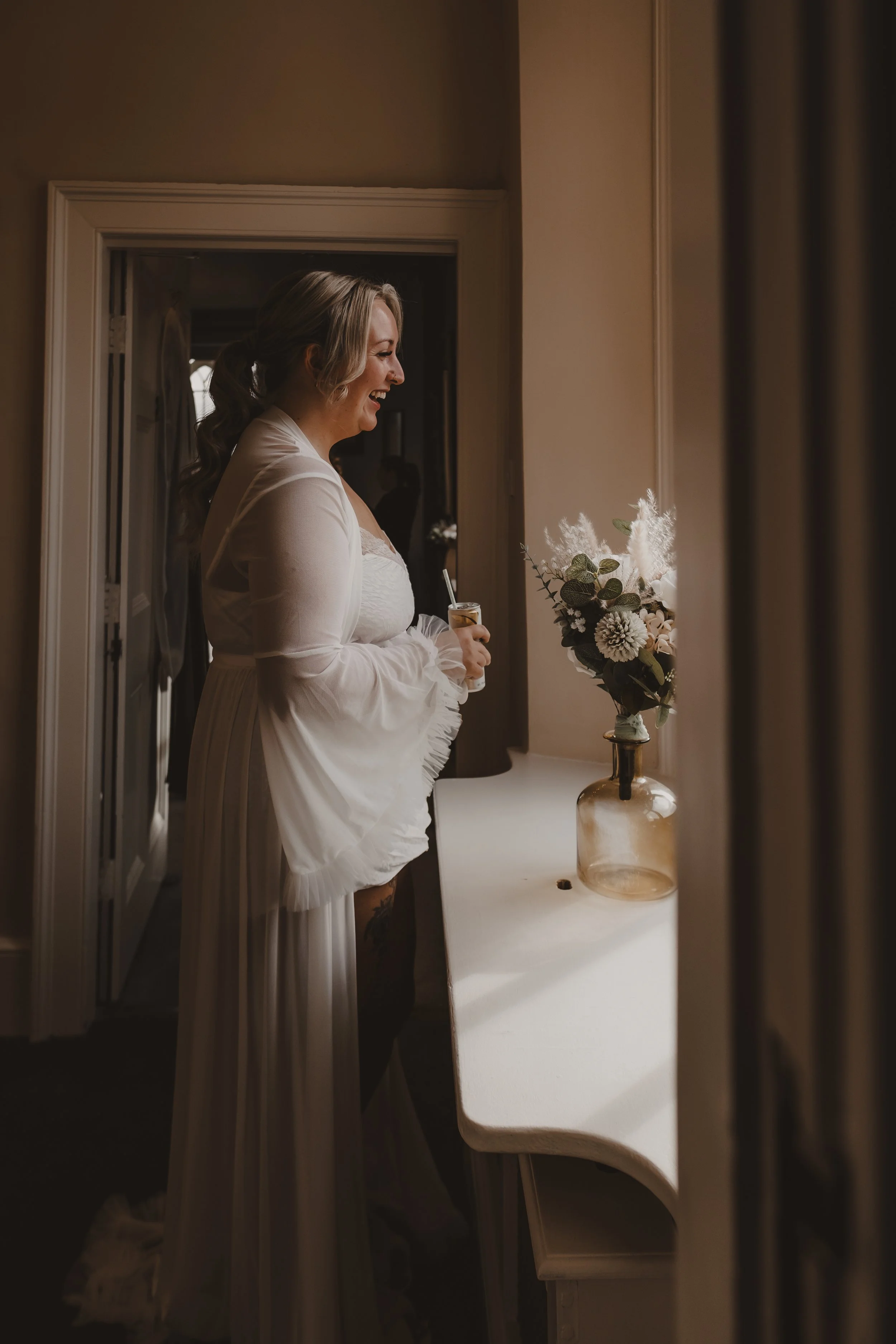 A woman in a white flowing bridal dressing gown standing by a window, holding a drink, smiling, with a vase of flowers on the table in front of her.