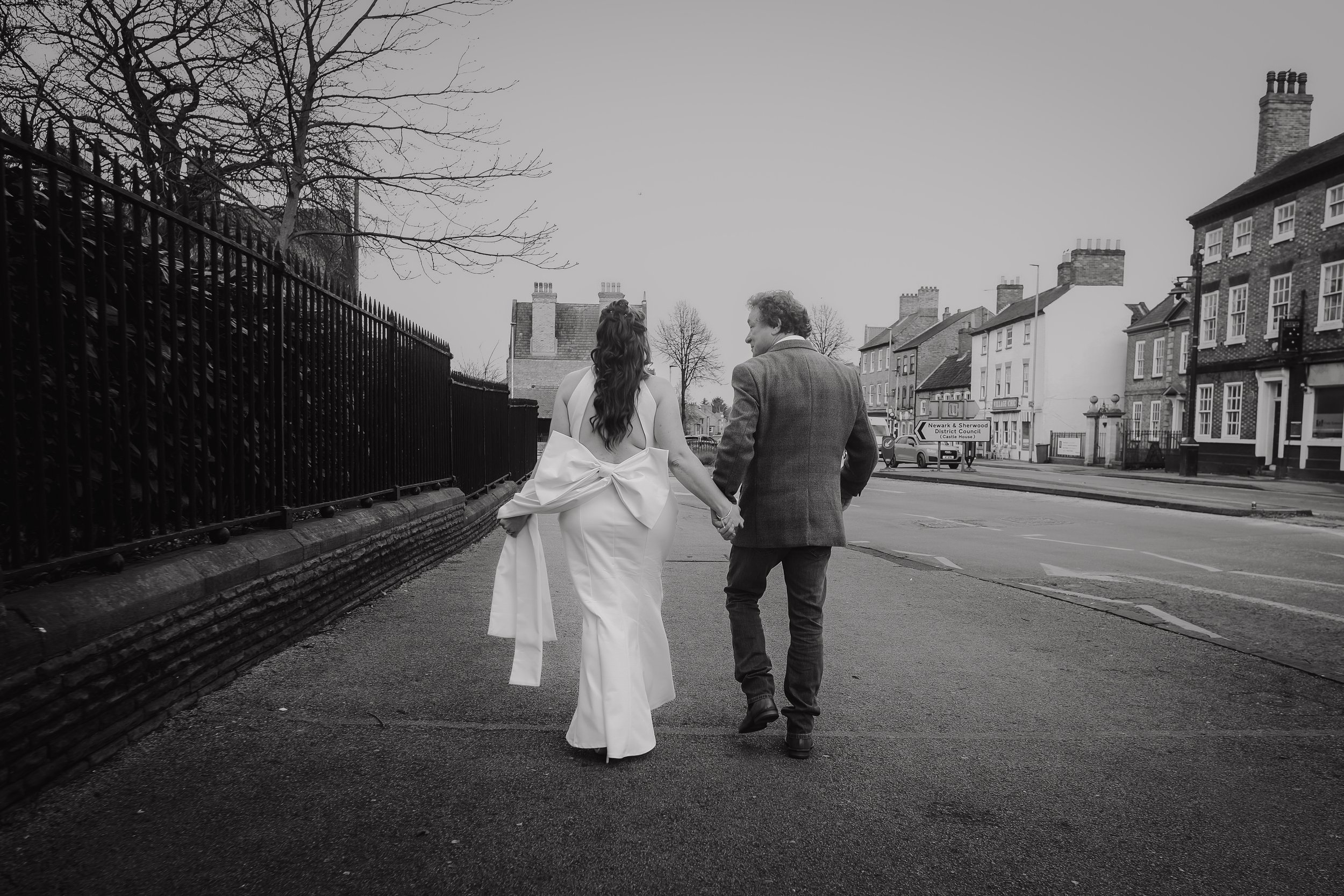 A black and white photo of a couple walking hand in hand on a city sidewalk, with the woman in a white dress with a large bow on the back, and the man in a suit jacket.