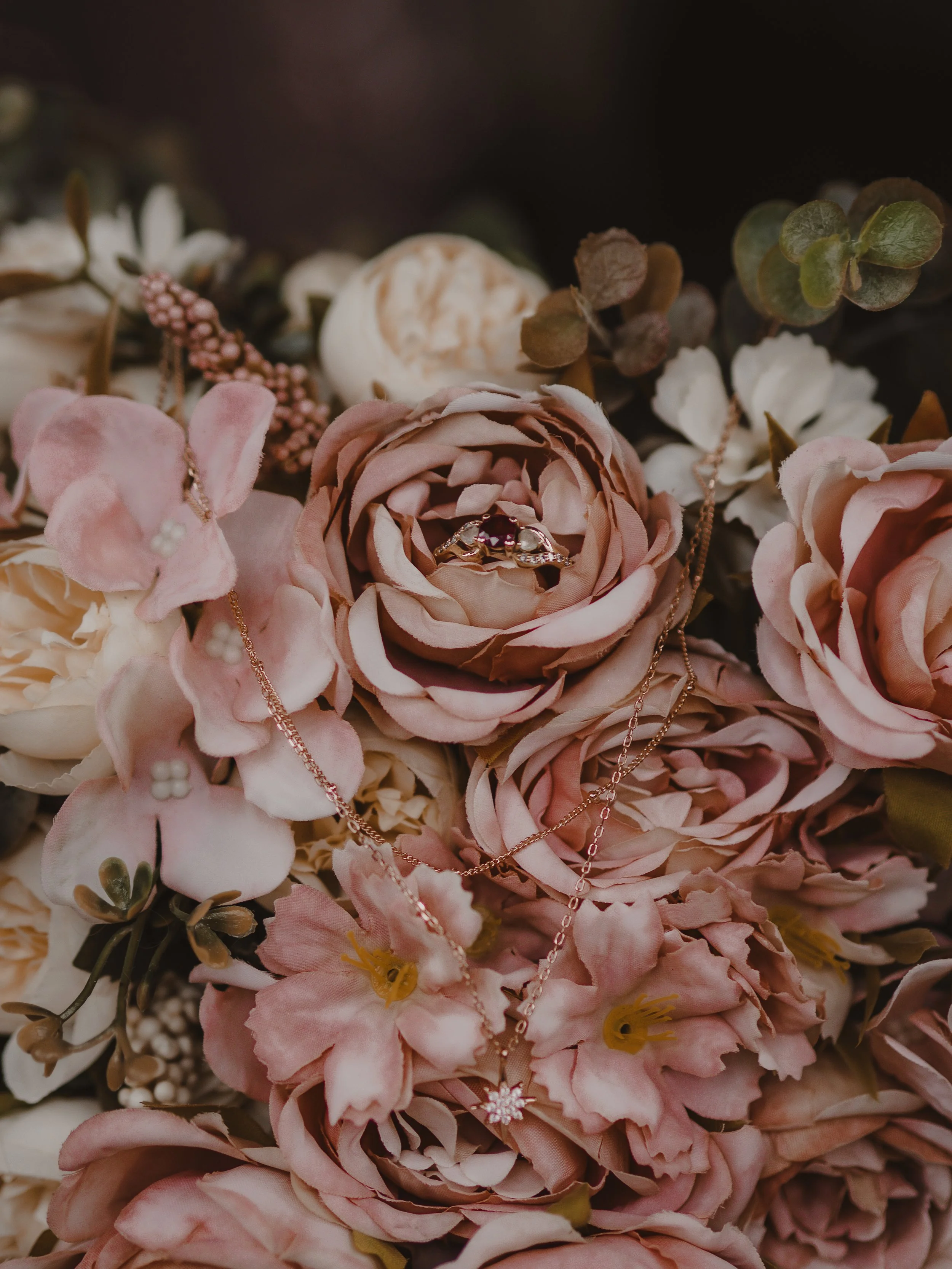 A close-up of a floral arrangement with pink and cream-colored flowers, featuring a ring and delicate necklaces resting on the flowers.