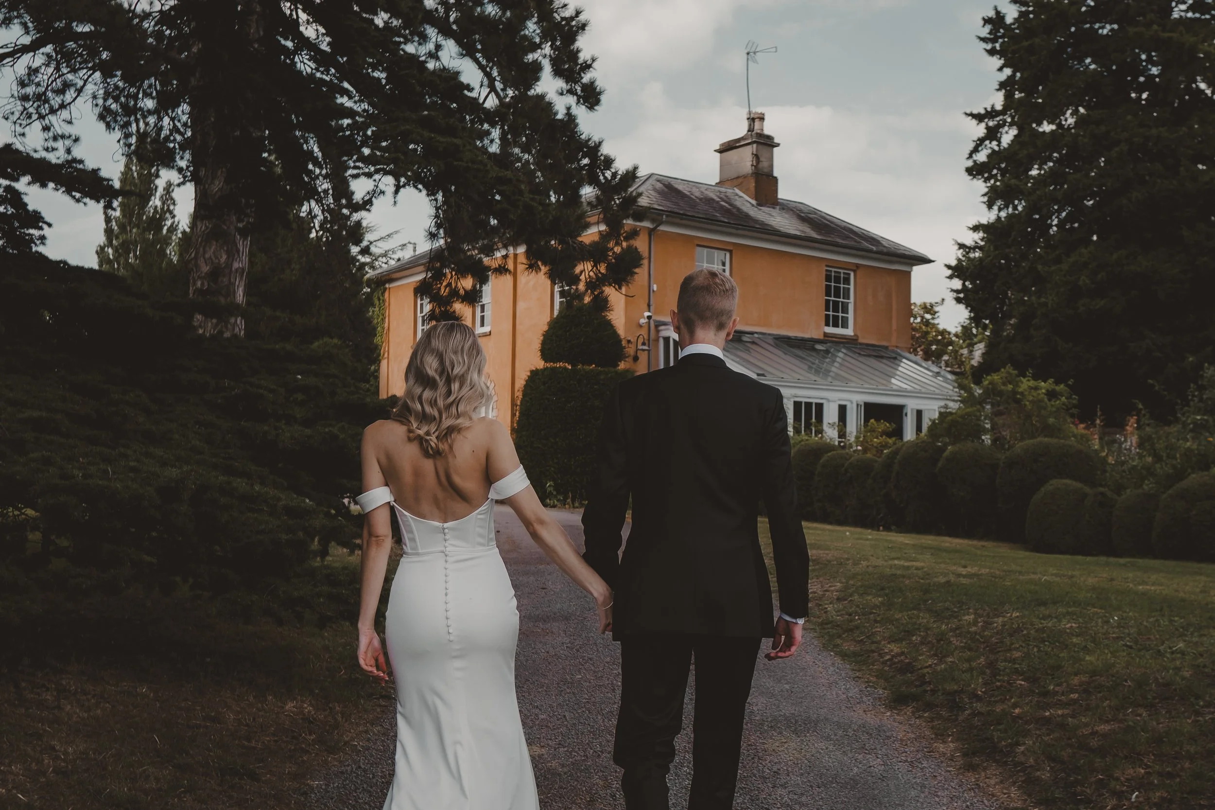 Couple dressed in wedding attire walking towards a large yellow house on a gravel path surrounded by greenery and trees.