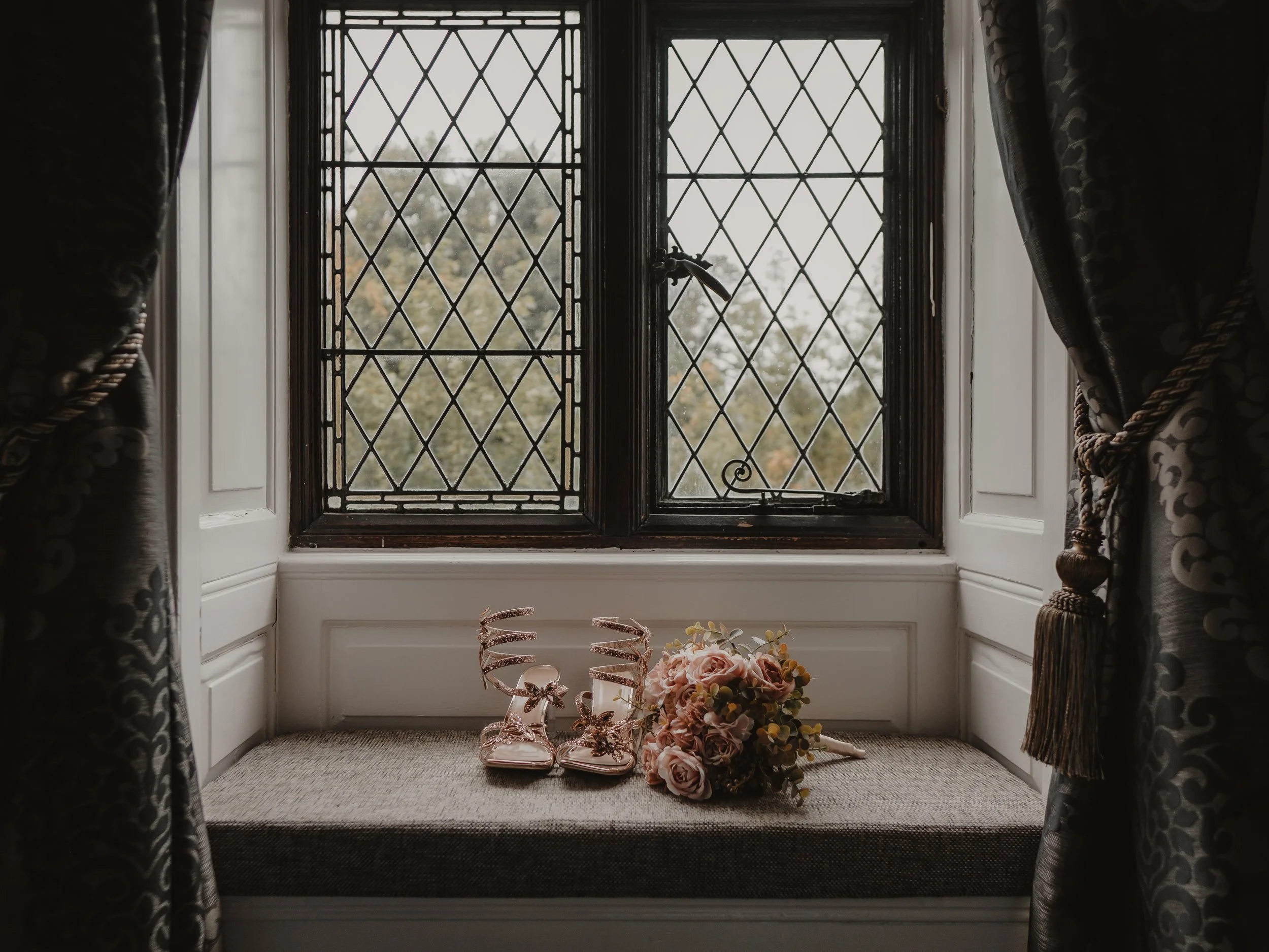 A windowsill with a pair of rose gold high-heeled shoes and a bouquet of pink and yellow flowers, framed by dark curtains with tassels.