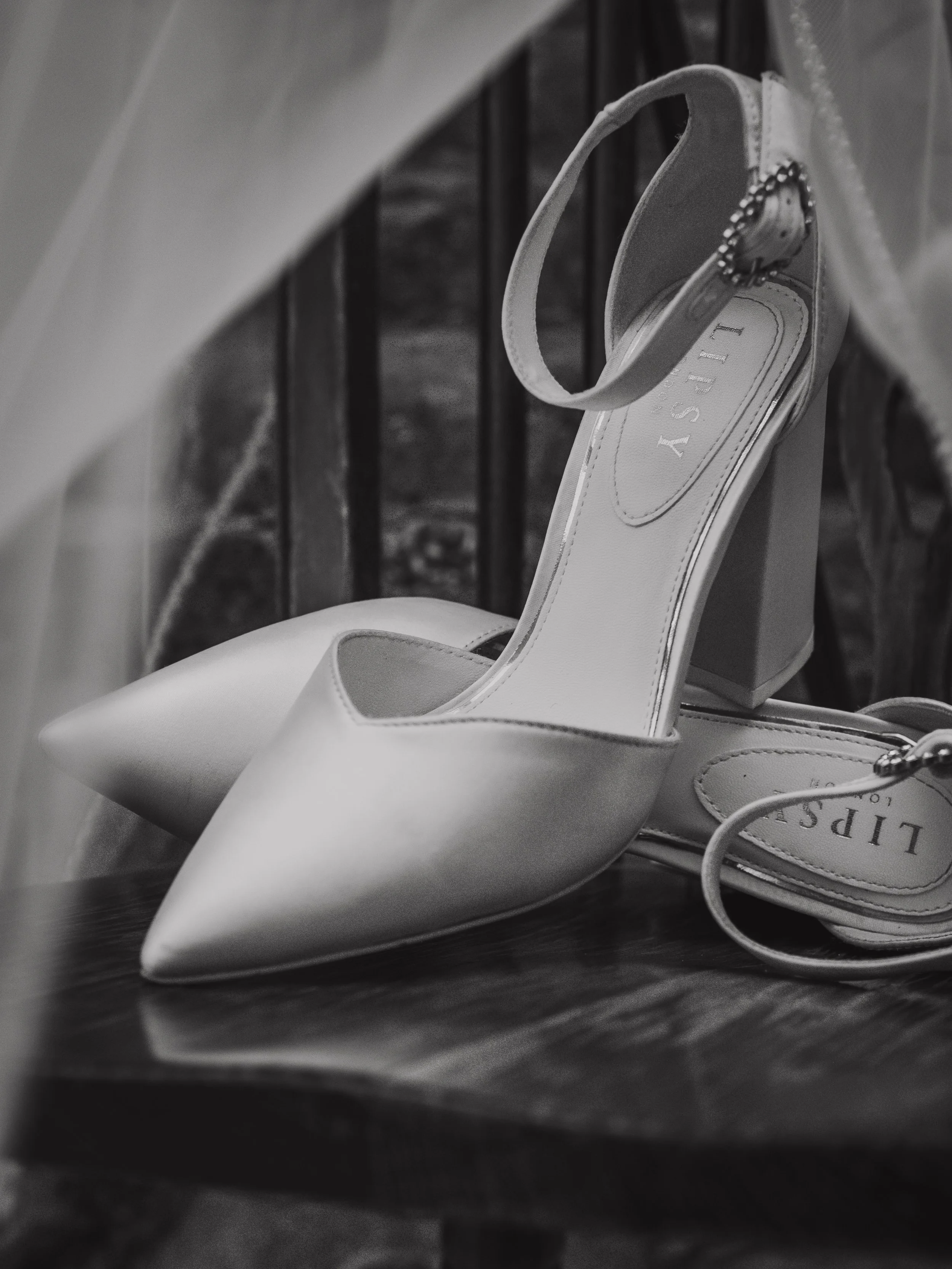 Black and white photo of a pair of white high-heeled shoes with pointed toes. The shoes have ankle straps and are placed on a wooden surface, leaning against a chair.