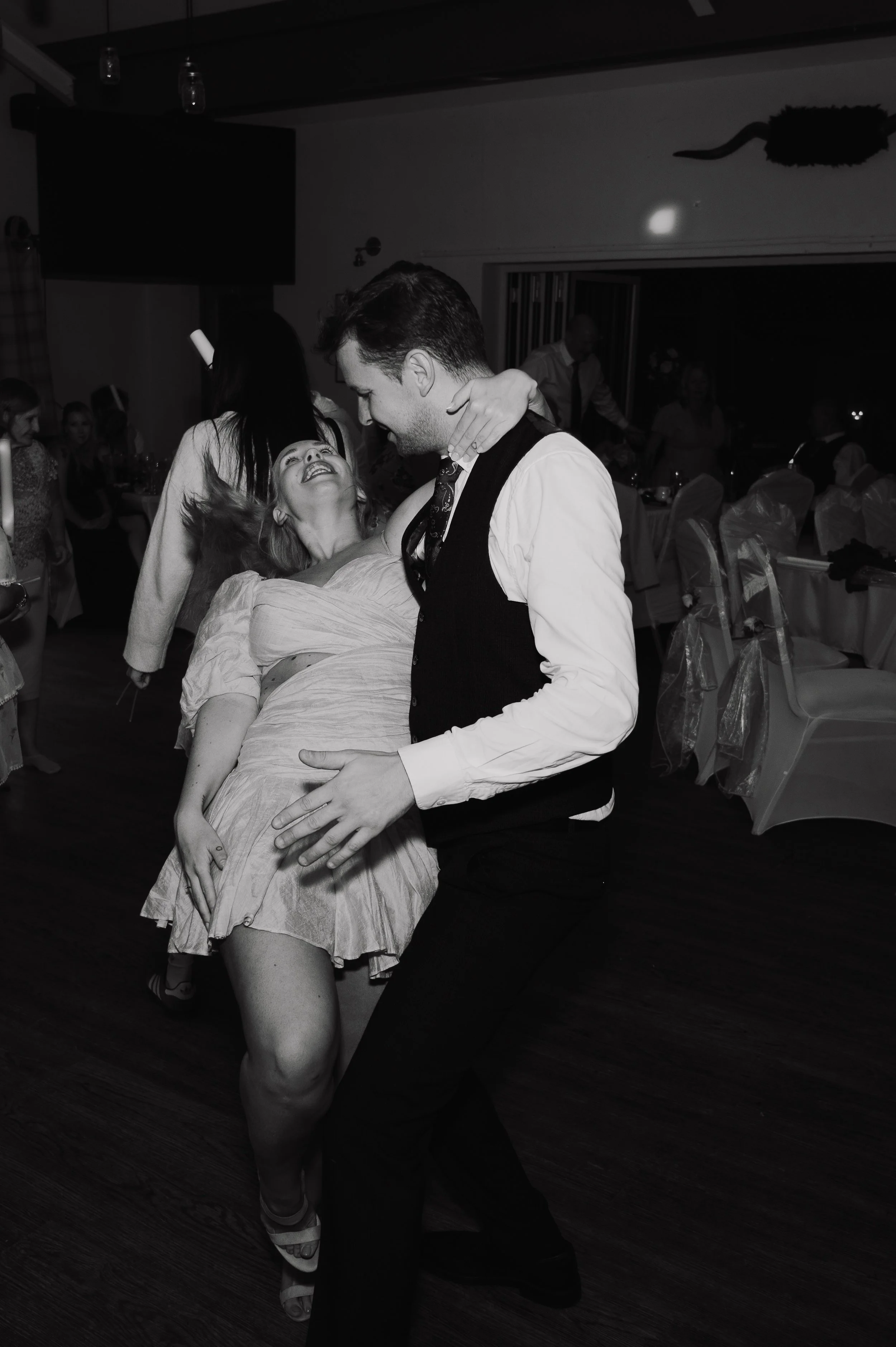A man and woman dance closely at a celebration, smiling and looking into each other's eyes, with other guests in the background.