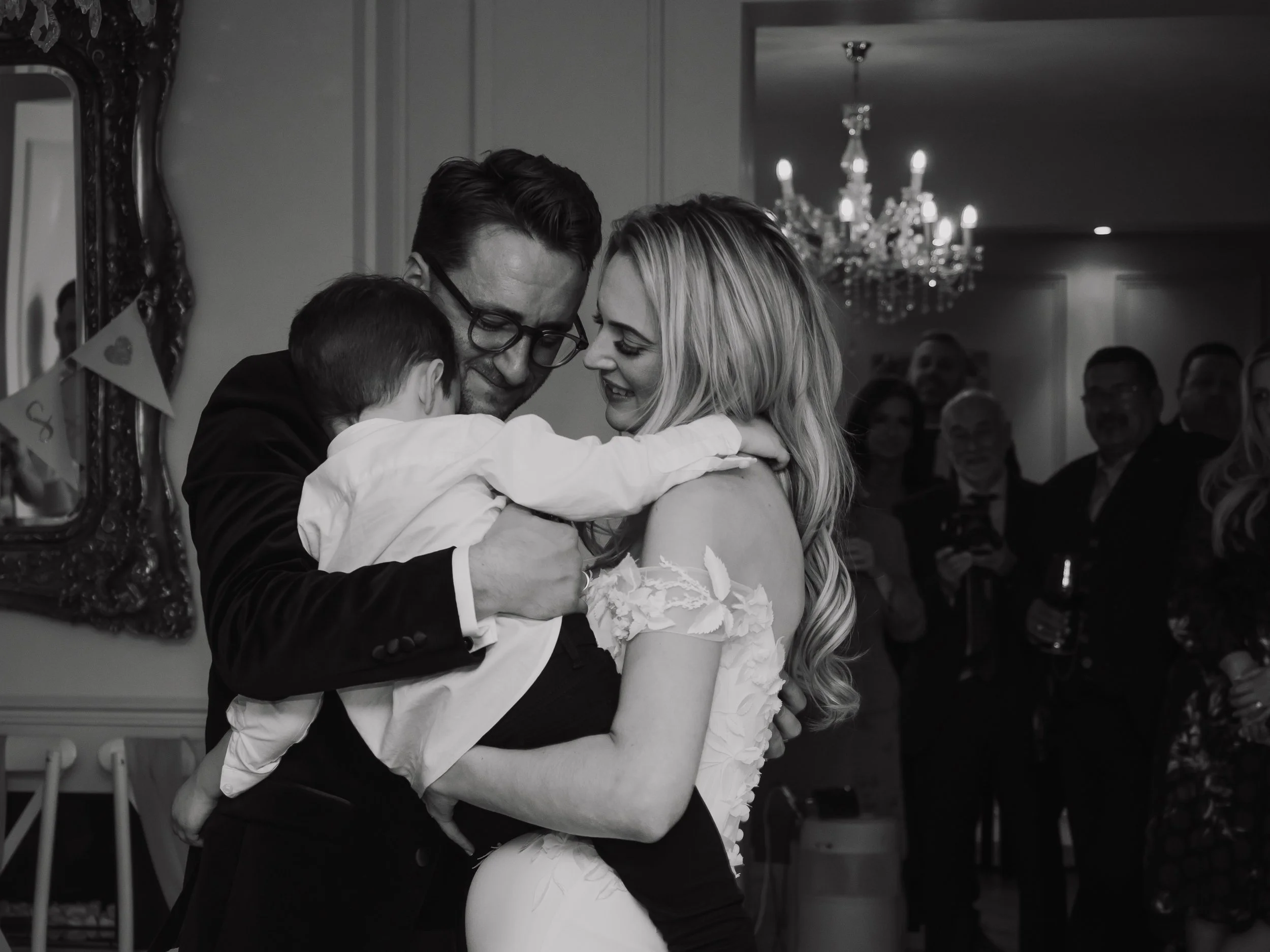 A family dancing at a wedding reception, including a bride, a groom, and a young child, with guests in the background.