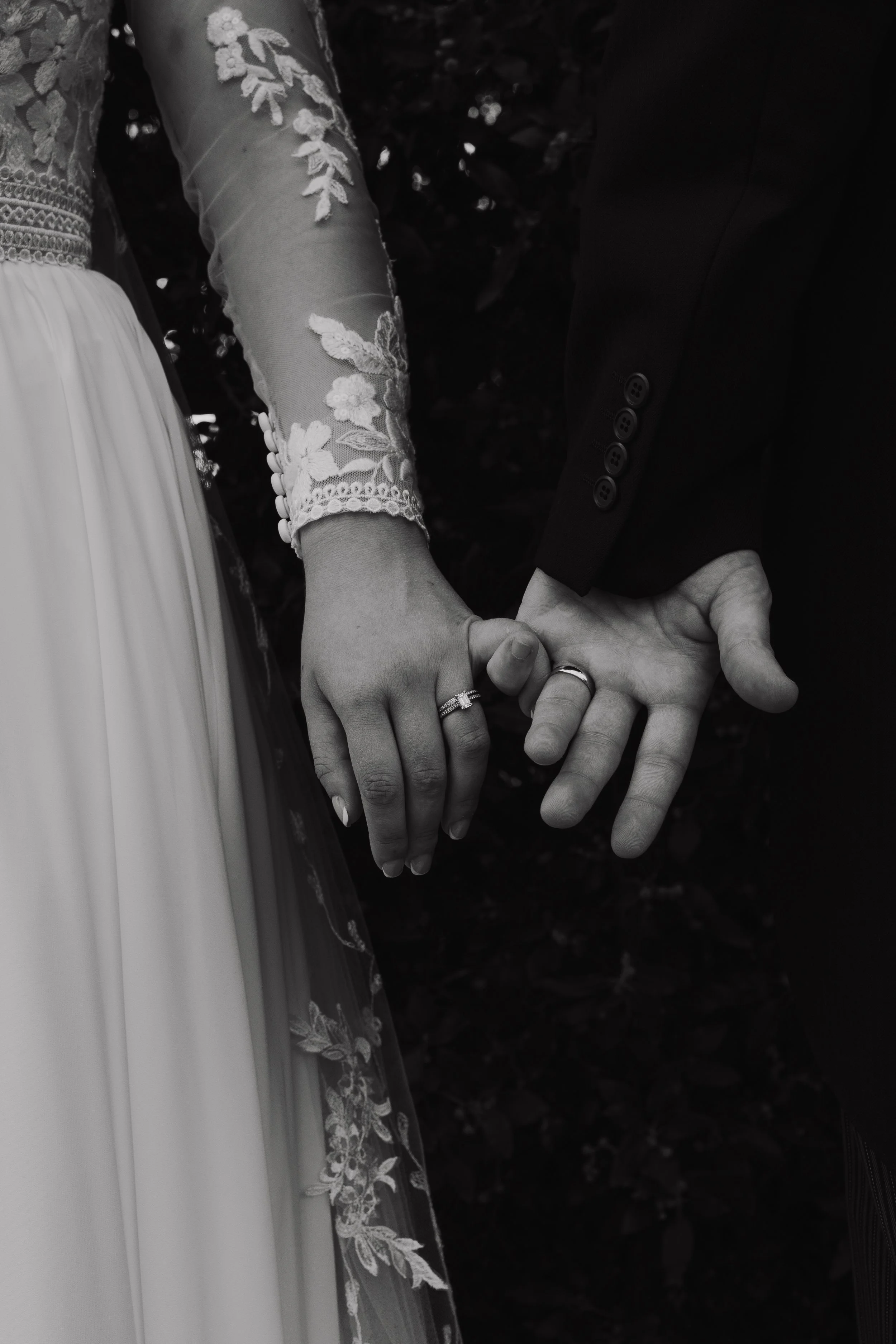 A black and white photo of a bride and groom holding hands, showing their wedding rings. The bride is wearing a lace long-sleeve wedding dress, and the groom is in a dark suit.